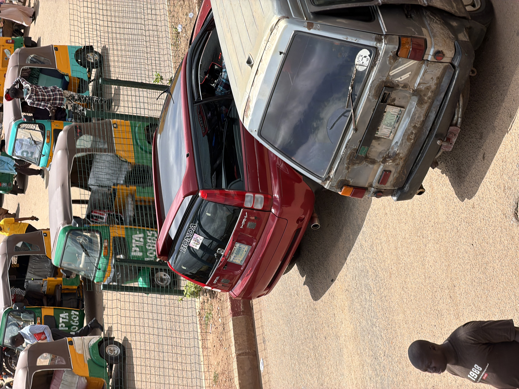 In the city of Bukuru, Nigeria, an urban scene unfolds with a focus on vehicular traffic and pedestrian activity. The foreground features a man in a brown t-shirt with "1988" printed on it, standing and looking to the left. Behind him, a weathered, light-colored van with visible rust and damage is parked, its rear bumper and license plate reading "JMT-300YR" are prominent. To the left of the van, a red Honda crossover SUV is parked, its rear window displaying a "HONDA" sticker and another smaller sticker. The vehicles are positioned on a dusty road or lot, with a chain-link fence running horizontally across the midground, separating the road from the background. Beyond the fence, several colorful auto-rickshaws, known locally as "Keke Napep," are visible. One yellow and green Keke is in motion, with a driver and passengers inside. Another is a yellow, green, and brown model, with a person in a checkered shirt standing near its open door. A third Keke, predominantly yellow and green, is also parked. A person in a yellow t-shirt and dark pants walks towards the right in the background. The overall environment suggests a busy roadside location with active transportation and commerce.