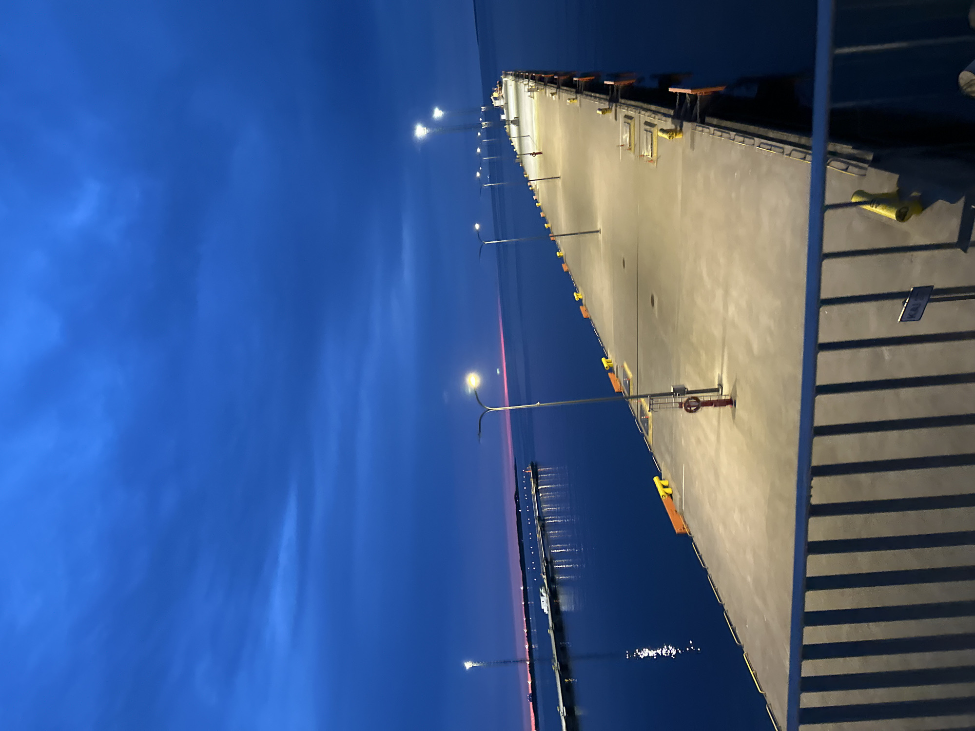 The image depicts a long, illuminated pier extending into a dark body of water under a twilight sky. Several streetlights cast bright pools of light along the concrete pier, which is lined with yellow bollards and orange fenders. A red fire hydrant is visible midway down the pier. In the distance, another pier with a tall tower and a white vehicle can be seen. The water reflects the lights from the pier and the distant horizon, which shows a faint pink and orange glow. A metal railing is in the foreground, casting long shadows. The scene appears to be a harbor or port area, possibly in Tallinn, Estonia, given the presence of such infrastructure. There are no people or active vessels in the frame.