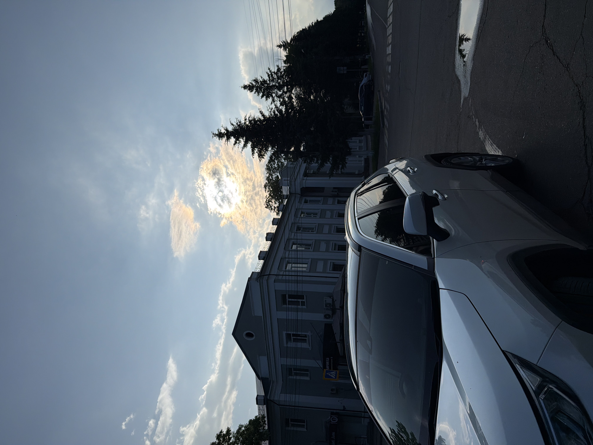 The image shows a white SUV parked on a paved road in Birobidzhan, Russia. In the background, a large, light-colored building with columns and multiple windows stands under a cloudy sky. Tall, dark green coniferous trees line the right side of the road, casting shadows. The sun is partially obscured by clouds, creating a bright halo effect. A puddle of water reflects the sky and trees on the road surface. The overall scene depicts an urban environment during daylight, possibly late afternoon or early evening, given the lighting conditions.