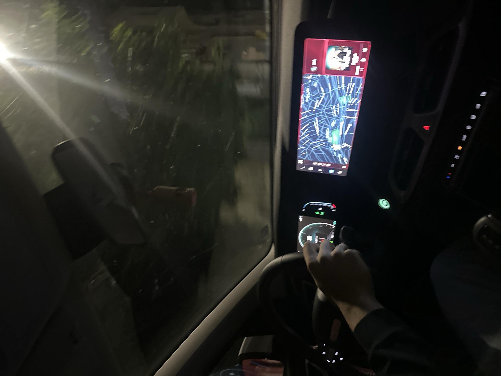 The image shows the interior of a car at night, possibly while driving. A person's hand is visible on the steering wheel, and the car's dashboard displays a map and music information. The scene is illuminated by the car's internal lights and the headlights of oncoming traffic or streetlights. The view through the windshield suggests a moderately lit urban environment with trees and buildings visible. The digital dashboard shows a navigation map with blue lines representing roads and a red indicator, likely for a song playing on a media player. The speedometer dial shows a speed of 30, possibly in kilometers per hour. There is no explicit information in the image that can definitively pinpoint the location to Shumen, Bulgaria.