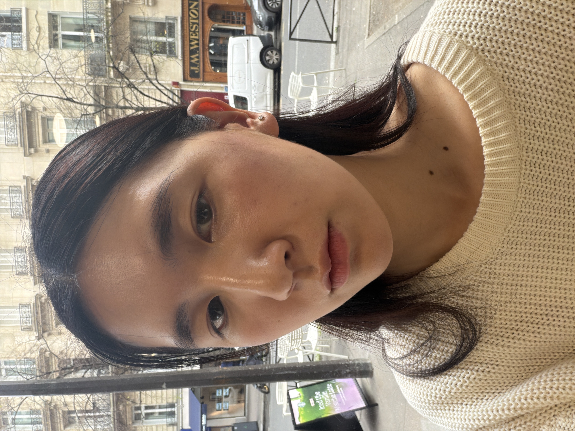 A close-up photograph depicts a person with dark hair and light skin wearing a cream-colored knit sweater. The person is facing the camera with a slight angle. The background reveals a Parisian street scene. Visible in the background are buildings with traditional Parisian architecture and a "J.M. Weston" storefront. A white van is also present. Other objects include what seems to be an advertisement for Iced vibe vanille and Matcha Latte. The image appears to have been taken outdoors during daylight.