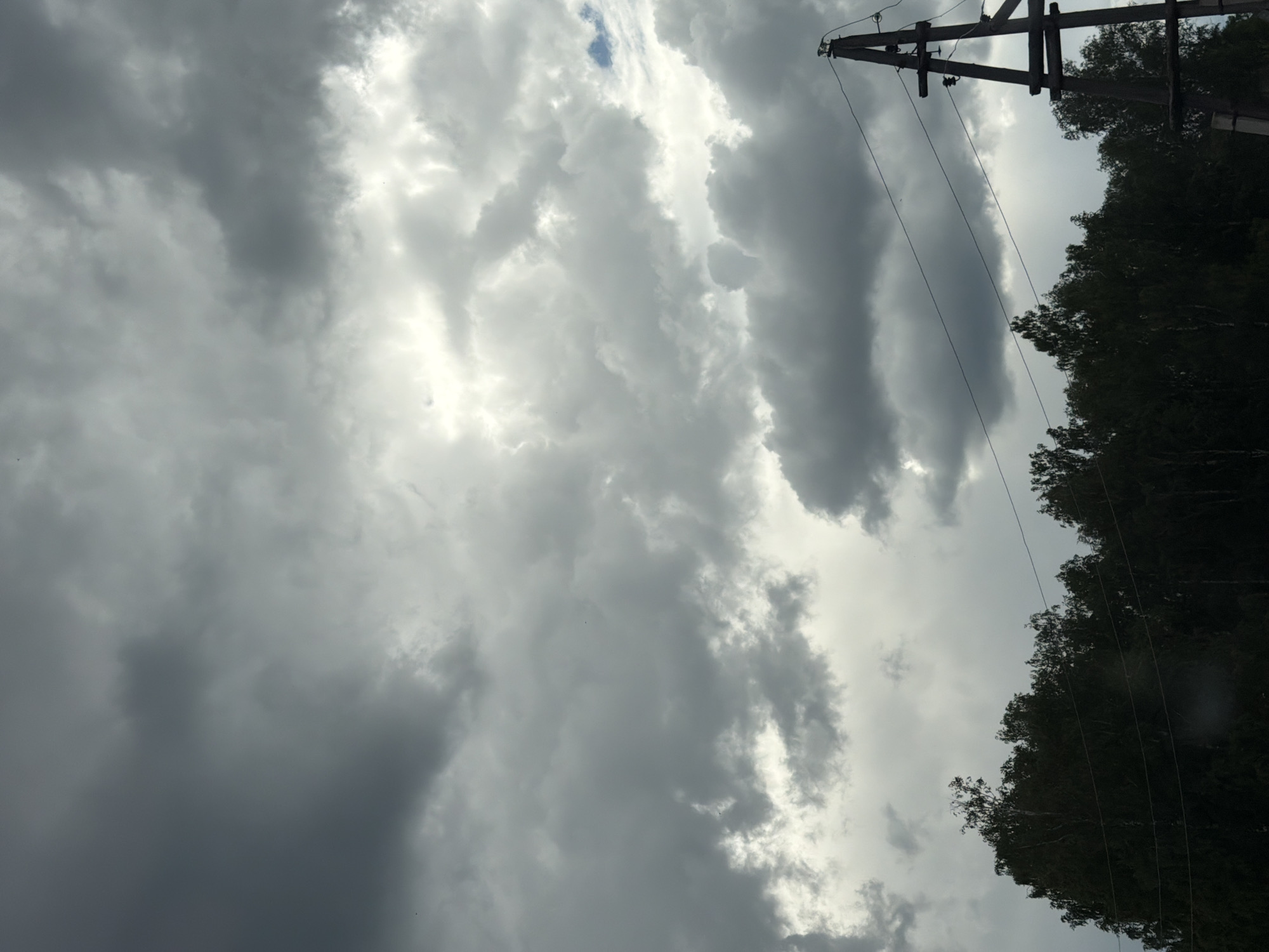 A landscape photograph captures a foreboding sky filled with dark, heavy clouds that dominate the upper half of the frame. A sliver of brighter sky peeks through the clouds in the upper center, suggesting a break in the overcast conditions. The lower portion of the image is dominated by the dark silhouettes of trees forming a dense line across the horizon. A wooden power pole with wires stretching across the scene stands prominently on the right side of the frame. The location is identified as Mezhevoy, Russia.