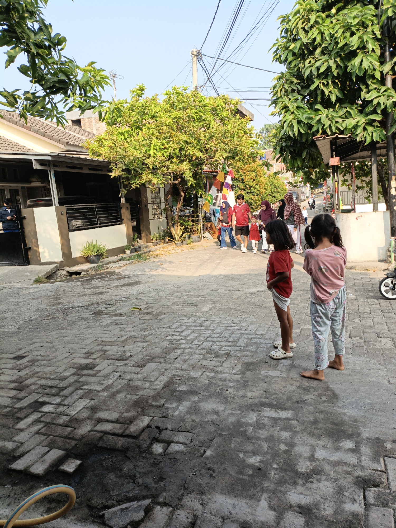 This is a full-shot, eye-level photograph taken in a residential street in Cilegon, Indonesia, under clear, bright daylight. The street is paved with interlocking gray bricks, some of which appear worn and wet. Two young girls are standing in the foreground, facing away from the camera and towards a group of people further down the street. The girl on the left wears a red t-shirt and white shorts with patterned clogs. The girl on the right wears a pink t-shirt with cartoon characters and patterned sweatpants.
In the background, a group of people are walking together. They are dressed in casual clothing, including a red t-shirt with black shorts, a hijab, and other varied attire. Some are carrying items, and there are flags or banners visible, suggesting a local event or celebration. The buildings lining the street are single-story homes with tiled roofs and covered porches. Lush green trees with dense foliage flank the street, casting shadows. Electrical wires crisscross overhead. A black car is parked in a driveway on the left side of the frame. A bicycle with a white rim is visible on the right side. The overall impression is a snapshot of everyday life in a suburban Indonesian neighborhood.