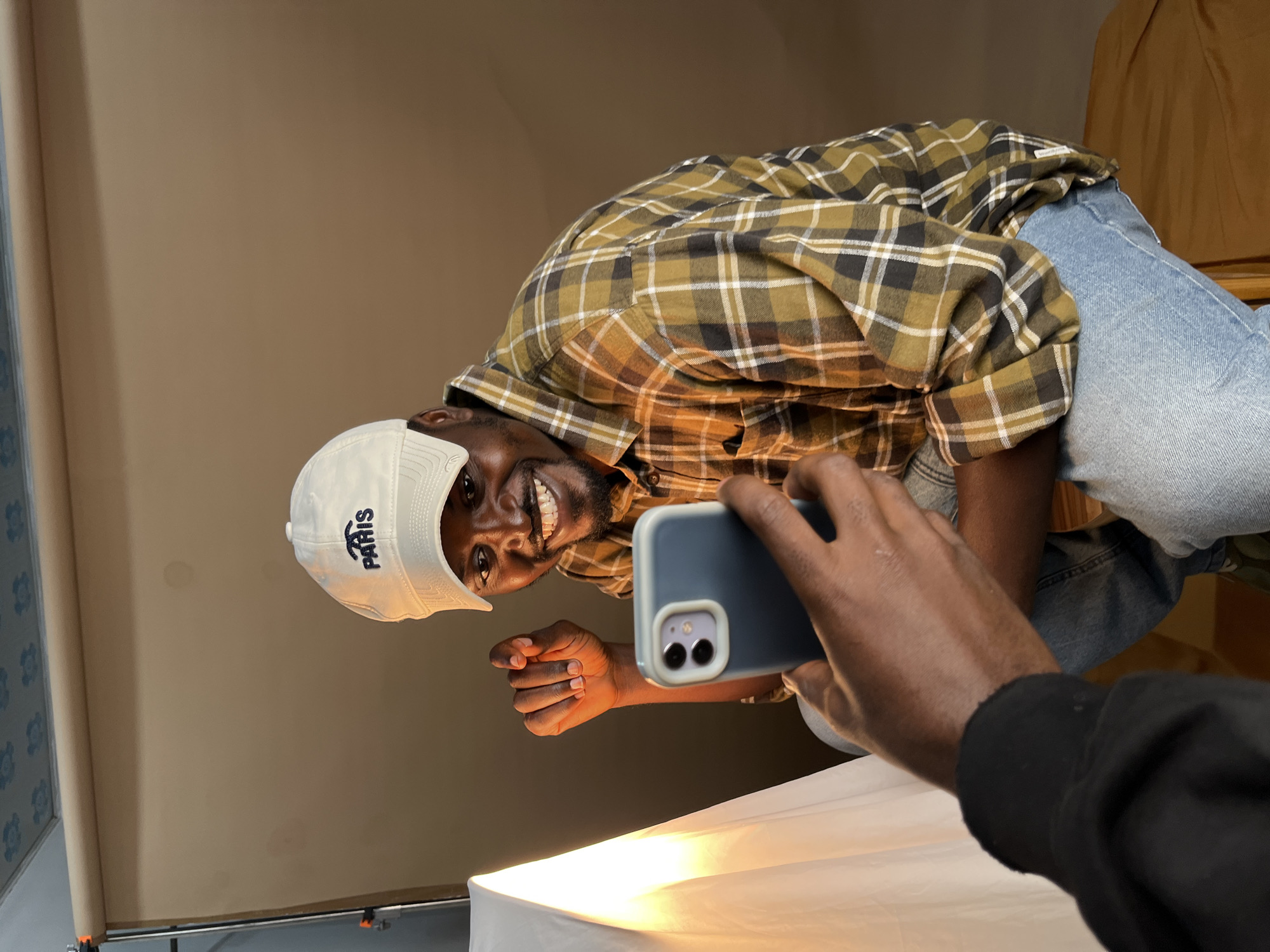 A man wearing a white cap with "PARIS" embroidered on it and a plaid shirt is posing for a picture being taken with a smartphone. The man is smiling broadly and making a fist with his left hand. He is sitting down, with his knees bent. To his left, a softbox illuminates the scene with a warm light. The background is a plain, neutral-colored studio backdrop. The image does not provide any information about a specific location in Gura Top, Nigeria.