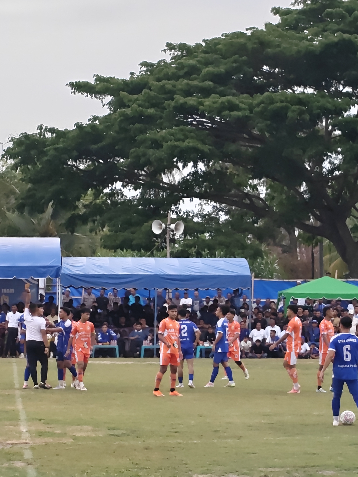 The image depicts an outdoor football match in Desa Meunasah Blang, Indonesia. A group of young men in orange and blue uniforms are on a green grassy field, some are actively playing while others observe. Spectators are visible in the background, seated under blue and green tents. The sky is overcast. A large tree with dense foliage dominates the upper portion of the image. There are speakers mounted on a pole to the right of the tents. The scene suggests a community sporting event taking place.