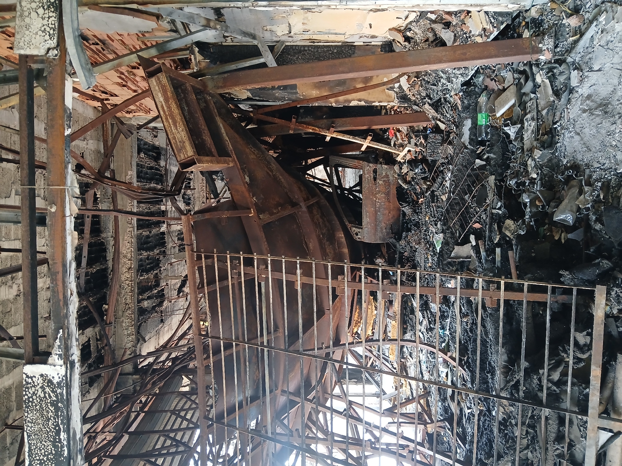 The image depicts the interior of a severely fire-damaged structure. Twisted and charred metal debris, including support beams and remnants of a staircase or railing, are scattered throughout the scene. Burned-out walls and remnants of a collapsed roof are visible. The devastation suggests a significant fire event. The location is not evident from the image alone. No people are present in the frame. The presence of a plastic bottle amongst the debris indicates post-fire activity. The overall environment is one of destruction and abandonment. The image implies a building collapse. Ciudad Guayana, Venezuela, if confirmed, would suggest a location facing urban decay.