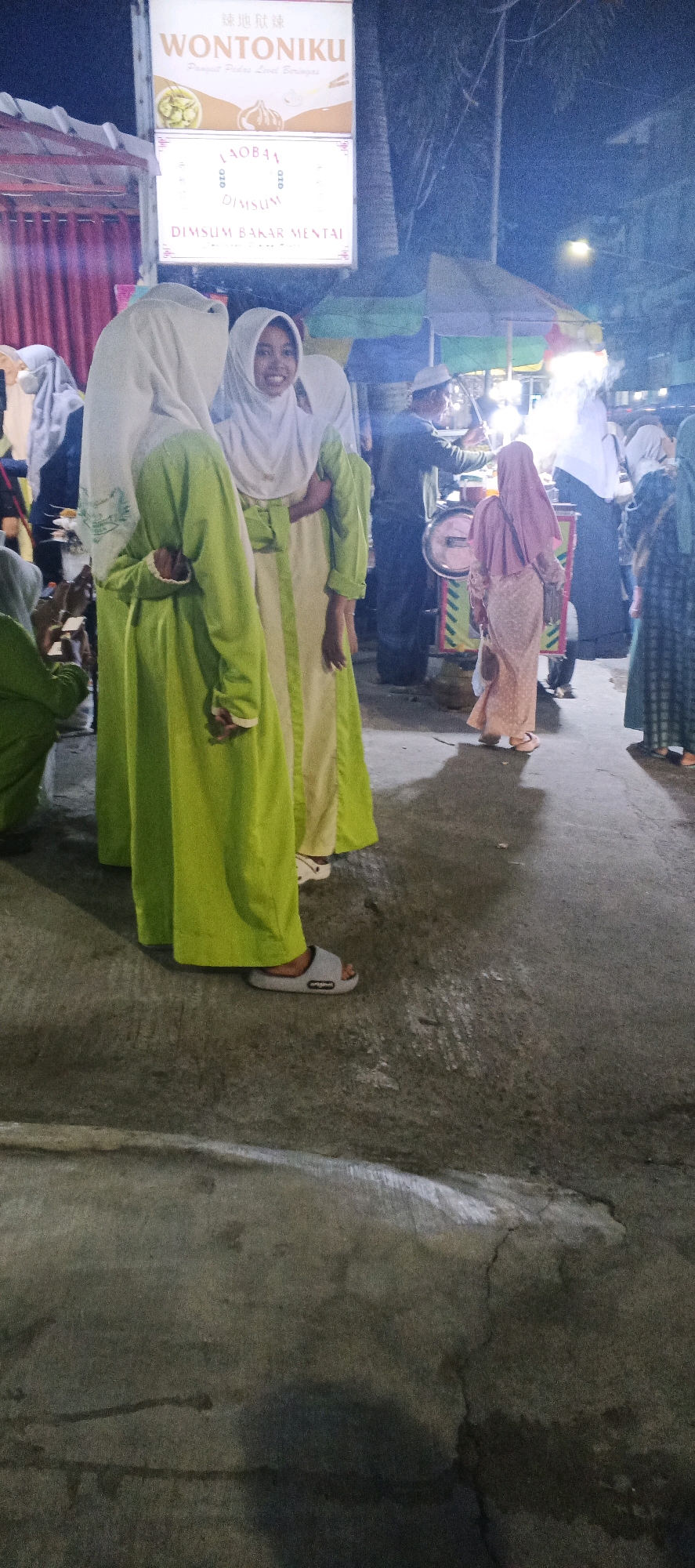 The image depicts a street scene at night in Sungelebak, Indonesia, possibly near a food stall. In the foreground, two individuals wearing light green and white hijabs and long dresses are standing. One is smiling and looking towards the camera. To the right, a person in a pink hijab and dress walks away from the camera, while behind them, a vendor in a dark blue shirt and white hat is operating a food cart. The food cart has a red circular object and appears to be illuminated. Signs are visible in the background, with one clearly displaying "WONTONIKU" in large letters, along with smaller text in Indonesian and what appears to be Chinese characters. The ground is paved with concrete.
