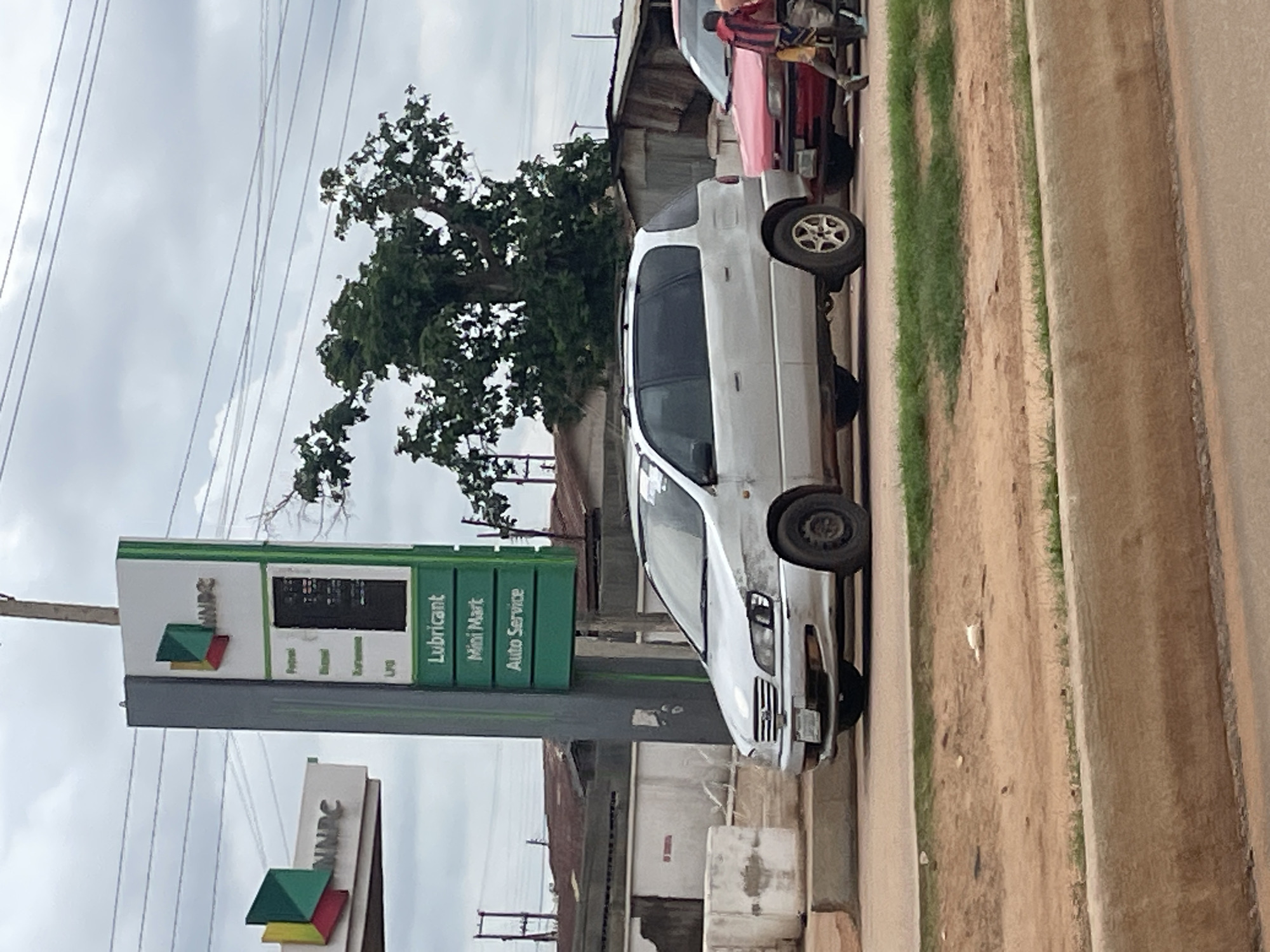 The image shows the NNPC (Nigerian National Petroleum Corporation) gas station in Minna, Nigeria. A silver minivan is parked in front of the station, to the left of a red car. A man and a child are standing next to the red car. The gas station has a tall green sign with "NNPC" at the top and listings for "Petrol," "Diesel," "Kerosene," "LPG," "Lubricant," "Mini Mart," and "Auto Service." The sky is overcast with power lines visible. The surrounding area appears to be a commercial or residential district with buildings and trees.