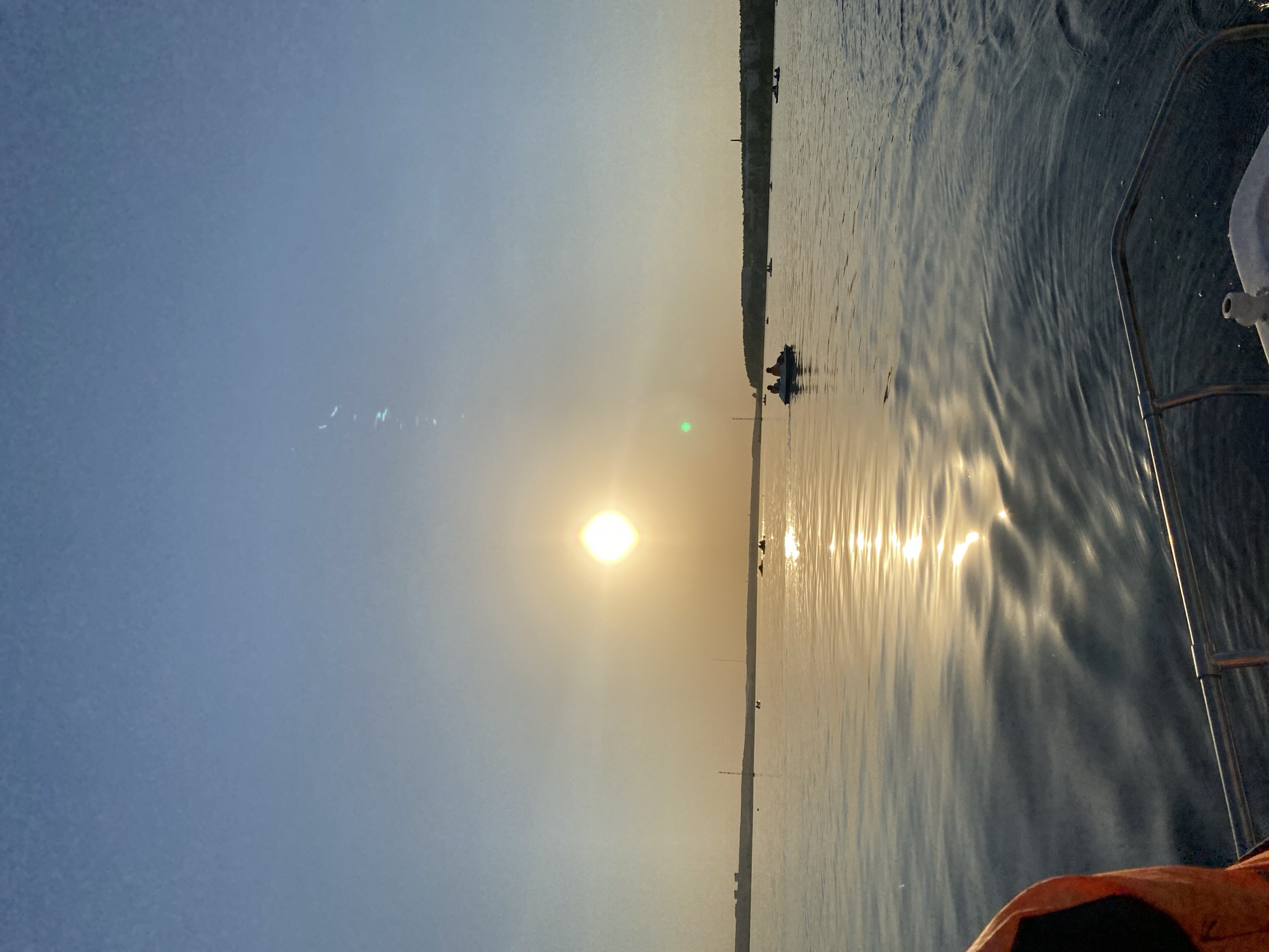 The image shows a sunset view over a body of water, likely a lake or reservoir, taken from a boat. The sun is positioned near the horizon, creating a bright glare reflected on the water's surface. The sky is a gradient of blue and yellow, indicating the time of day. In the distance, a shoreline is visible with some vegetation. There are a few small boats or watercraft on the water. The foreground includes a portion of the boat the photo was taken from, including part of the railing and what appears to be a life vest.
Based on the scene details, particularly the large body of water and the relatively flat terrain, the location is likely the Izhevsk Reservoir in Izhevsk, Russia. The Izhevsk Reservoir is a prominent feature of the city and is often used for recreation and water activities. The photo captures a typical view of the sunset over the reservoir, with boats and the shoreline visible in the distance.