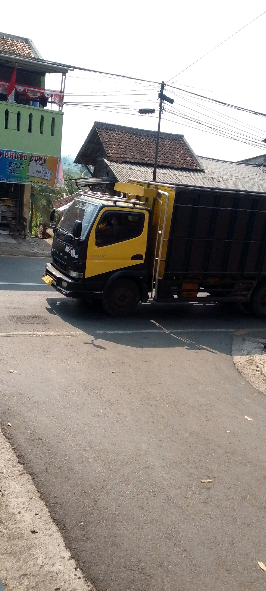 A yellow and black FUSO truck is traveling on a paved road in Cilaku, Indonesia. The truck has a black cargo box. In the background, there are several buildings with tiled roofs, including a green building with "PHOTO COPY" signage. There are also utility poles with many wires visible against a bright, overcast sky. The road surface is asphalt and appears to be in good condition. The truck is the primary subject of the image.