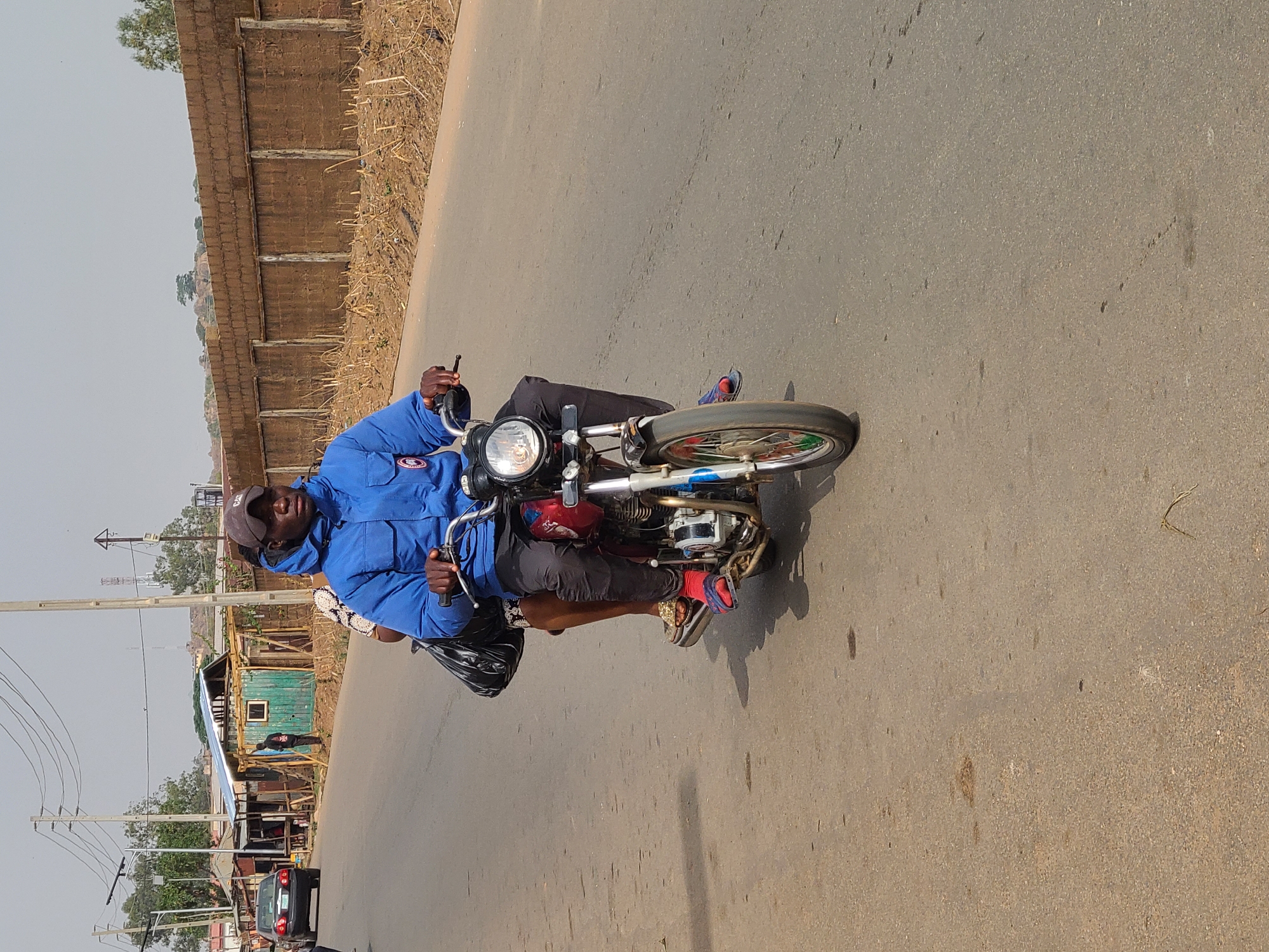 The image shows a man riding a red motorcycle on an asphalt road in Barakin Lerawi, Nigeria. The man is wearing a blue jacket, black pants, a cap, and red and brown sandals. He has a passenger on the back of the motorcycle. In the background, there is a low brick wall on the left side and buildings with corrugated metal roofs and wooden structures on the right side. Power lines and utility poles are visible in the distance. A black car is parked on the side of the road. The sky is overcast.