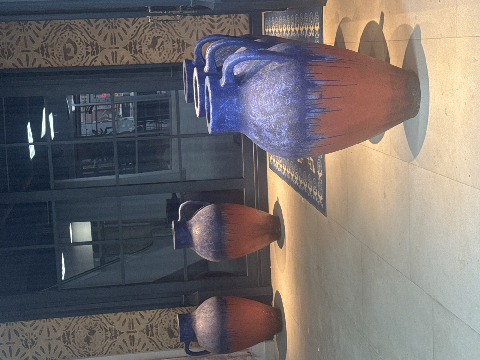 Four large, decorative ceramic vases with blue tops and reddish-orange bottoms are displayed in what appears to be an indoor setting. The vases have a glazed finish with drips running down from the blue section onto the orange. Two vases are positioned to the left, and three are lined up to the right, with the farthest right vase partially obscured. They are situated on a light-colored tiled floor, with a patterned rug visible beneath the vases on the right. Behind the vases is a large glass-paned door, reflecting interior lighting and structures, and textured wallpaper adorns the wall to the left and right of the door. The overall lighting is dim with warm tones cast on the floor. There is no indication that this scene is located in Ufa, Russia.