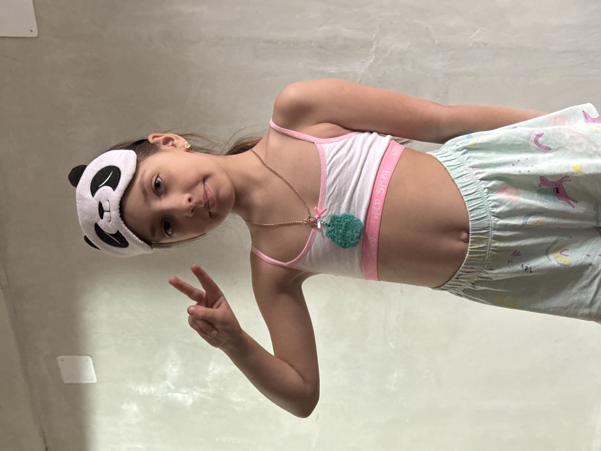 A young girl is depicted in a full-body, vertically oriented indoor photograph, taken from a low angle in Votkinsk, Russia. Her head is tilted, and she gazes directly at the camera with a subtle smile. She makes a peace sign with her right hand.
The girl wears light sleepwear: a white and pink sports bra or crop top with the words "WILD SPIRIT" visible on the pink elastic band, and light blue or teal pajama shorts adorned with colorful patterns that appear to be whimsical shapes, possibly unicorns, rainbows, or stars. A panda-themed sleep mask is pushed up onto her forehead. She accessorizes with a gold-colored necklace featuring a small, fluffy green charm, and a small gold earring in her right ear.
The background consists of a plain, light-colored wall, likely off-white or light gray, with a rectangular white object, possibly a light switch or outlet cover, visible on the left side. The lighting is bright and even, indicating an indoor setting. The casual attire and sleep mask suggest a relaxed, possibly morning or evening, atmosphere.
