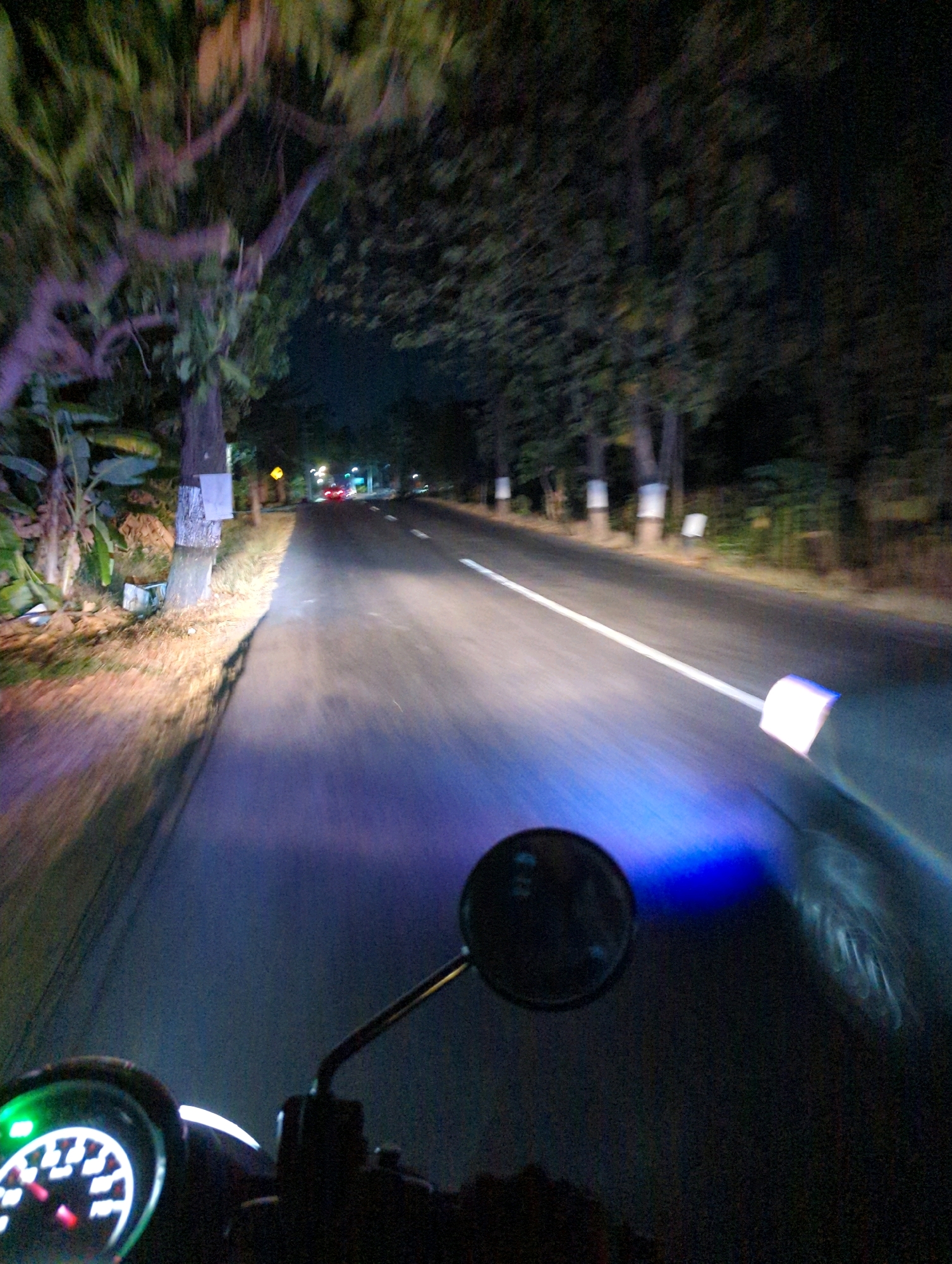 The image depicts a first-person perspective of riding a motorcycle at night on a road in Kacangan, Indonesia. The road is paved and has a white line marking the center. Trees line both sides of the road, with some illuminated by artificial lights. The motorcycle's headlight casts a blue glow on the road ahead. A portion of the motorcycle's dashboard, including the speedometer, is visible in the foreground. There are a few cars visible in the distance, their headlights and taillights illuminated. The overall scene suggests a nighttime journey through a rural or semi-rural area of Kacangan.