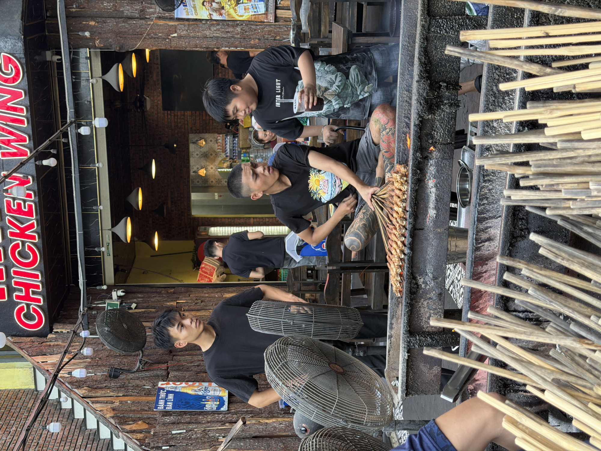 This media file captures a lively street food scene in Da Nang, Vietnam, likely during the daytime given the bright lighting. The primary subjects are several young men involved in preparing and serving food. In the foreground, a man with extensive tattoos on his leg and arm is holding a large skewer of grilled meat, possibly chicken. He is wearing a black t-shirt with a tropical graphic. Behind him, another young man in a black t-shirt with the words "INTER LIGHT" printed on it, is holding a cup with a straw and looking towards the food being prepared. A third individual, also in a black t-shirt, is visible further in the background, appearing to work near a food stall.
The setting is an outdoor food stall with a rustic wooden facade. Several large industrial fans are strategically placed, suggesting a warm climate. Above the food preparation area, a prominent red neon sign spells out "CHICKEN WING", clearly indicating the specialty of the establishment. The grilling area itself is dark and appears to be actively used, with visible embers and smoke. Piles of wooden skewers, some already laden with food and others waiting to be used, are a significant visual element.
The overall mood seems casual and industrious, typical of a busy food service environment. The focus is on the preparation and presentation of grilled skewers. There is also a sign with Vietnamese text, "ĐÁNH THỨC BẦN LINH", and an advertisement for Tiger beer. The presence of the fans and the outdoor setting imply a warm, possibly humid, weather condition.