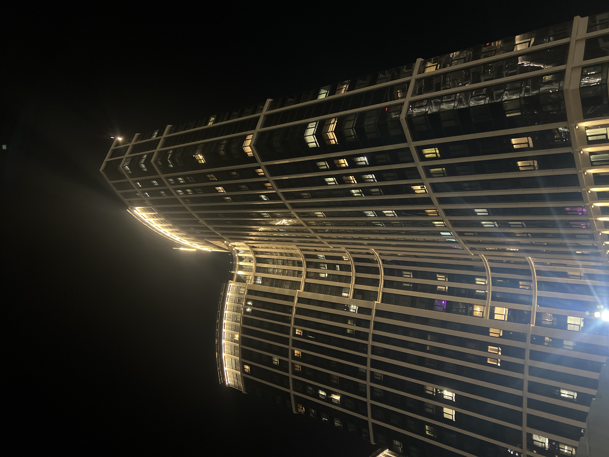 The image depicts a tall, modern skyscraper at night. The building has a curved facade with a grid-like structure of white lines and dark glass. Lights are visible in some of the windows. The location appears to be Moscow, Russia. The surrounding environment is dark.
