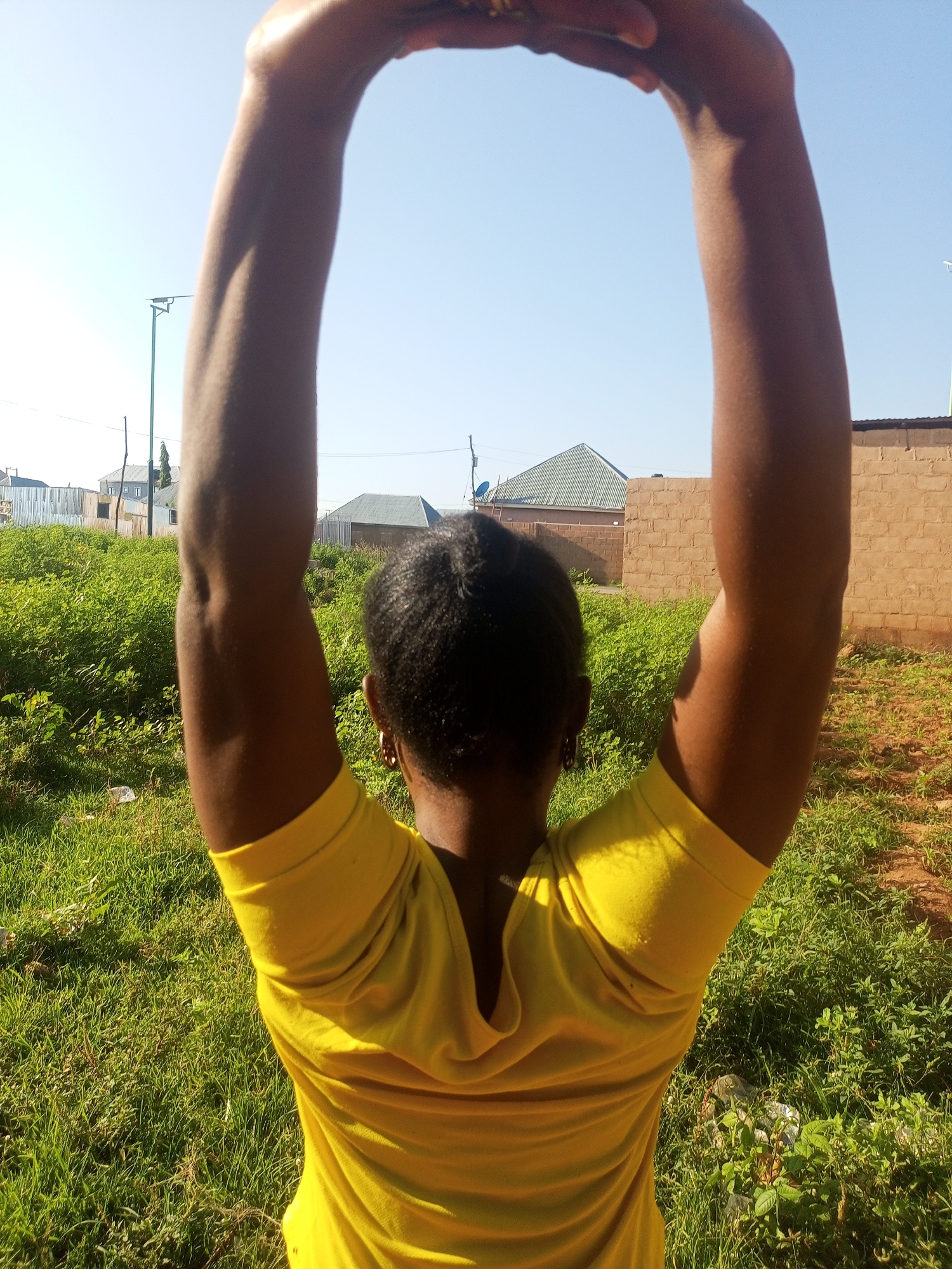 An overhead, close-up rear view shows a person standing outdoors, raising their arms above their head to form a circle. The person is wearing a bright yellow t-shirt. The background consists of a clear blue sky, green grass, and buildings in the distance. The setting appears to be a rural or semi-urban area in Tempure, Nigeria, with brick buildings and corrugated metal roofs visible. The lighting suggests it is daytime, likely morning or afternoon.
