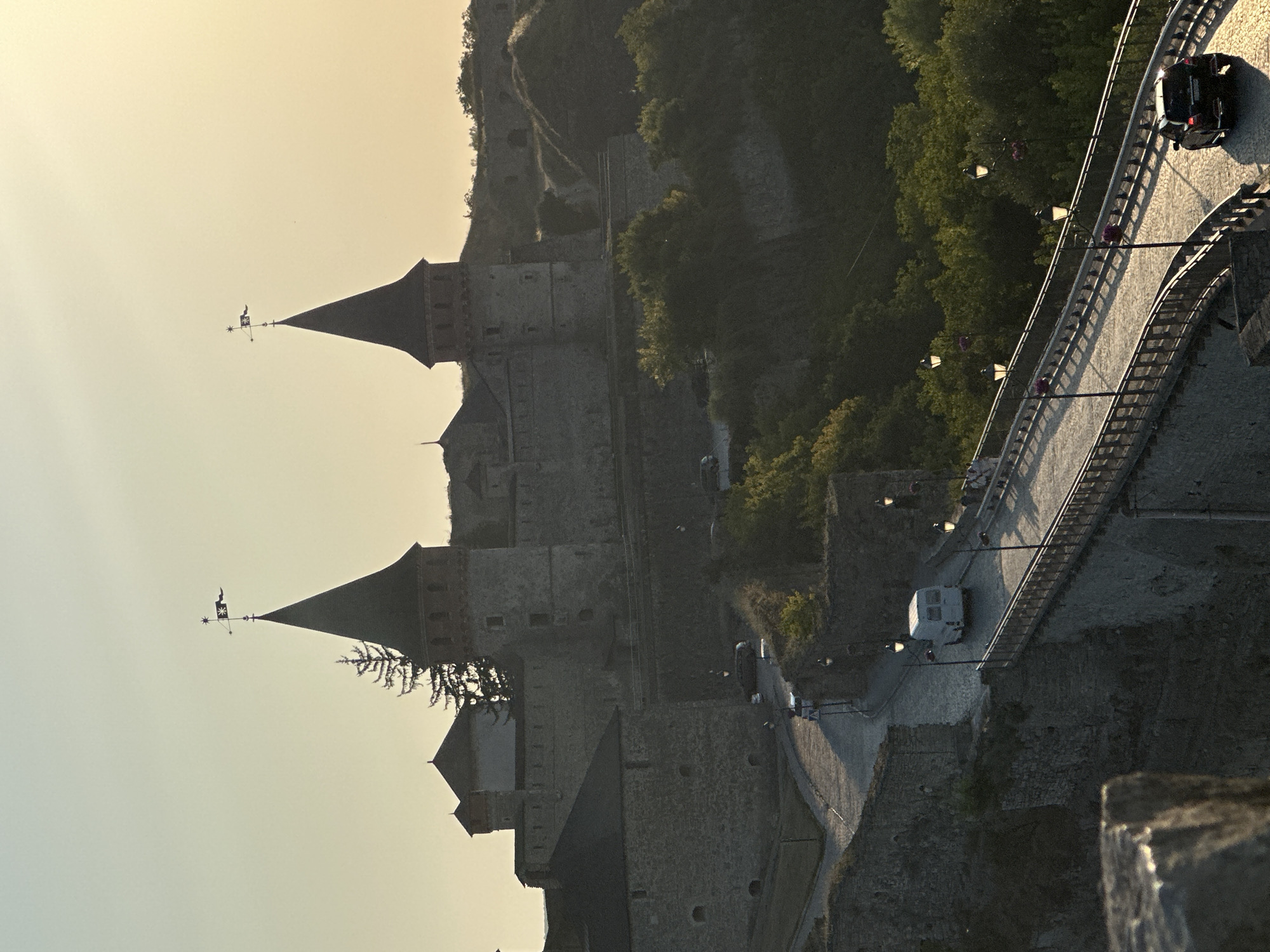 The image shows the Kamianets-Podilskyi Castle in Kamianets-Podilskyi, Ukraine, at sunset. Two tall, conical towers with flags stand prominently against a pale yellow sky. The castle walls are made of stone, and there are other buildings within the complex. A cobblestone road winds up towards the castle, and a bridge with decorative lampposts spans a valley. A white van is driving on the road, and a black car is on the bridge. There are some trees and vegetation surrounding the castle and on the slopes. The scene is bathed in the warm, soft light of the setting sun.