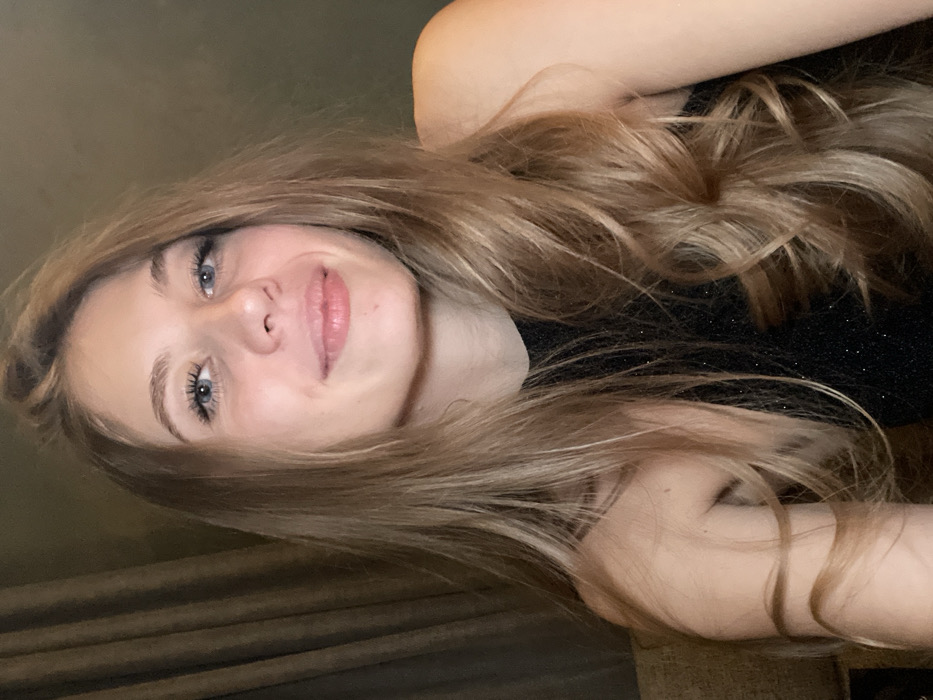 A young woman with fair skin, blue eyes, and light brown hair is featured. Her hair is styled in loose waves. She is wearing a dark, possibly sparkling, top. The background includes a beige-colored curtain or draped fabric. The photograph is taken at a slight angle. Her lips are slightly parted, suggesting a calm expression.