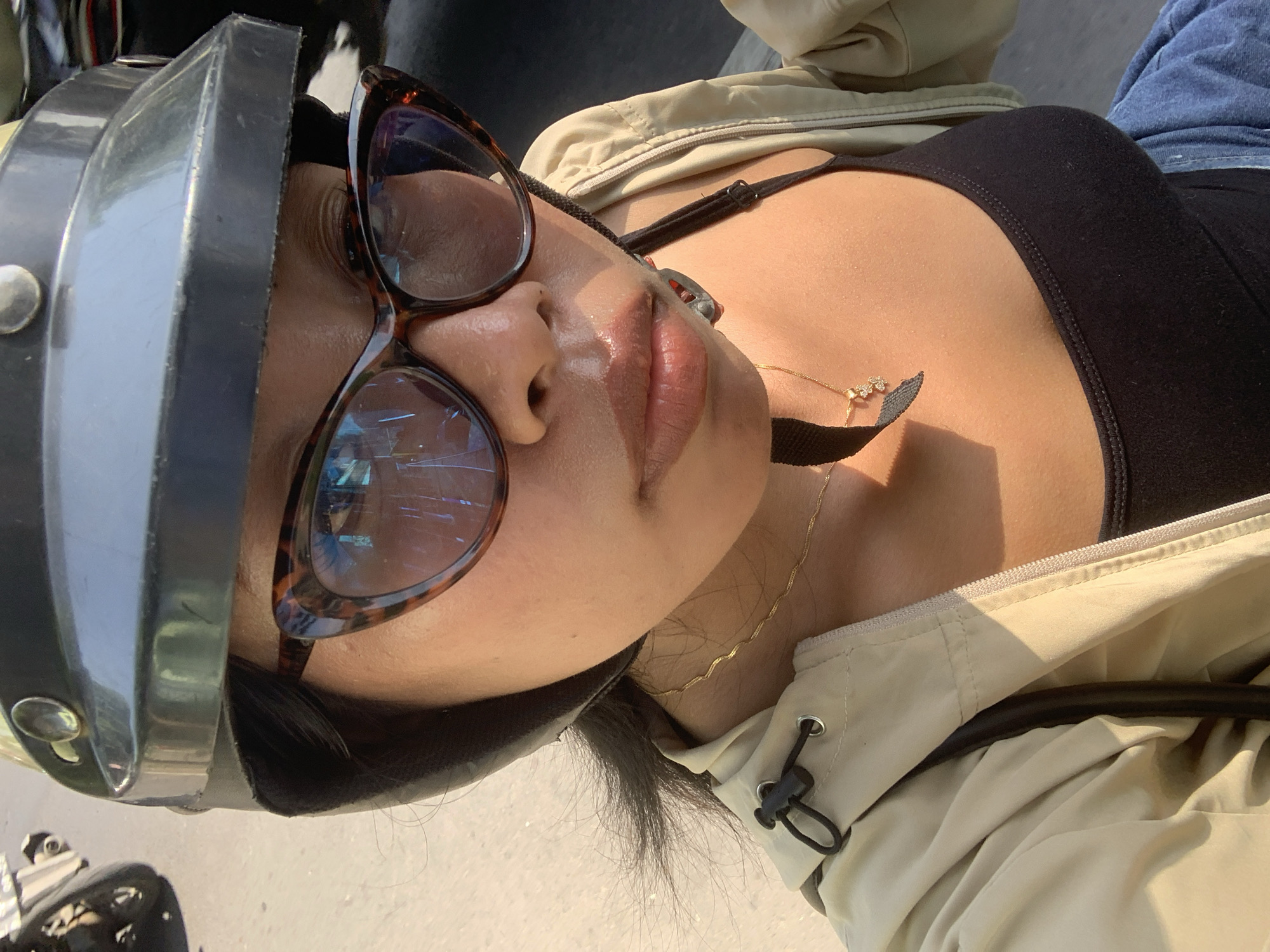 A woman wearing a black helmet and tortoiseshell-patterned glasses is pictured from the chest up, looking forward. She has dark hair and is wearing a beige jacket over a black tank top. A delicate gold necklace with a small pendant is visible around her neck. The background is blurred but suggests an outdoor setting, possibly a street, with hints of a motorcycle wheel on the left and a dark, indistinct background on the right. The lighting indicates it may be daytime. The image does not contain information to identify the location as Tamanbaru, Indonesia.