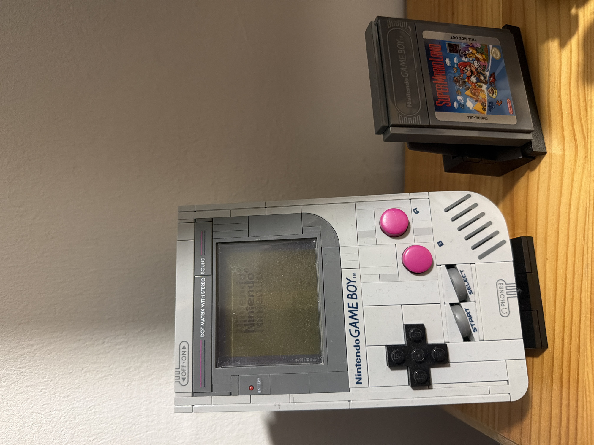 A Nintendo Game Boy and a Super Mario Land cartridge are displayed on a wooden surface. The Game Boy is a model made of Lego bricks, with a grey and white color scheme. The screen shows a faded "Nintendo" logo. The Super Mario Land cartridge is in a black plastic stand, and the label depicts Mario and other characters from the game. The background is a textured light-colored wall. The location is not specified.