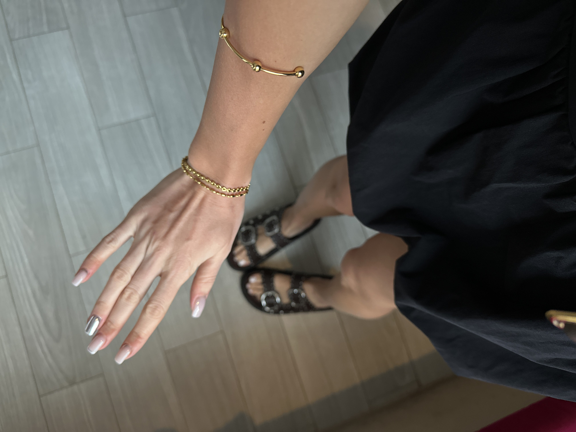 A high-angle medium shot shows a person with light skin wearing a short black dress and dark brown sandals with silver buckles. The person is standing on a wooden floor with light and dark grain patterns. The person's left arm is extended towards the left of the frame. The person has two gold bracelets on their left arm: a thin chain bracelet at the wrist and an open cuff bracelet higher up. The person's fingernails are painted nude and white with one silver accent nail. The location is Unknown, Indonesia.