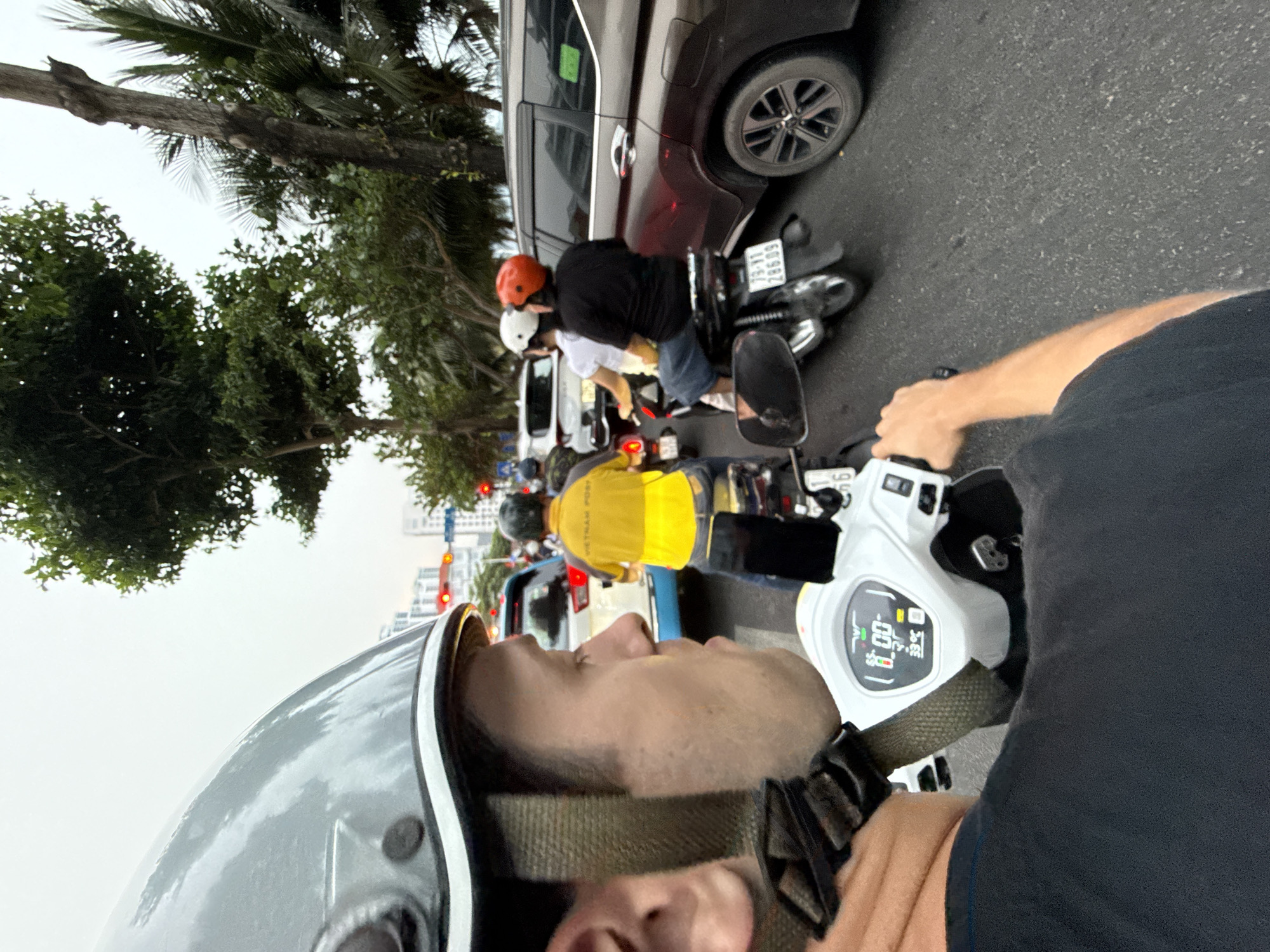 The image captures a first-person perspective from someone riding a white scooter in traffic in Bac Nha Trang Ward, Vietnam. The sky above is overcast or cloudy.
In the foreground, part of the photographer's face, a grey and white helmet, a black t-shirt, and an arm holding the scooter's handlebar are visible. The scooter's digital dashboard is clearly displayed, showing "00" (likely speed), a battery icon, and the time "13:34".
Ahead, a line of motorcycles and scooters is stopped in traffic. Directly in front, a person wearing a black t-shirt, shorts, and an orange helmet is on a motorcycle. Behind them, a dark gray SUV is also stopped. Further ahead in the lane, another individual on a scooter wears a bright yellow vest with "VIETNAM POST" written on it and a black helmet. A red traffic light and buildings are visible in the background, suggesting an urban setting. Lush green trees line the left side of the street. A motorcycle license plate "73-B1 266.09" is visible behind the SUV. The scene depicts a typical daytime traffic congestion.