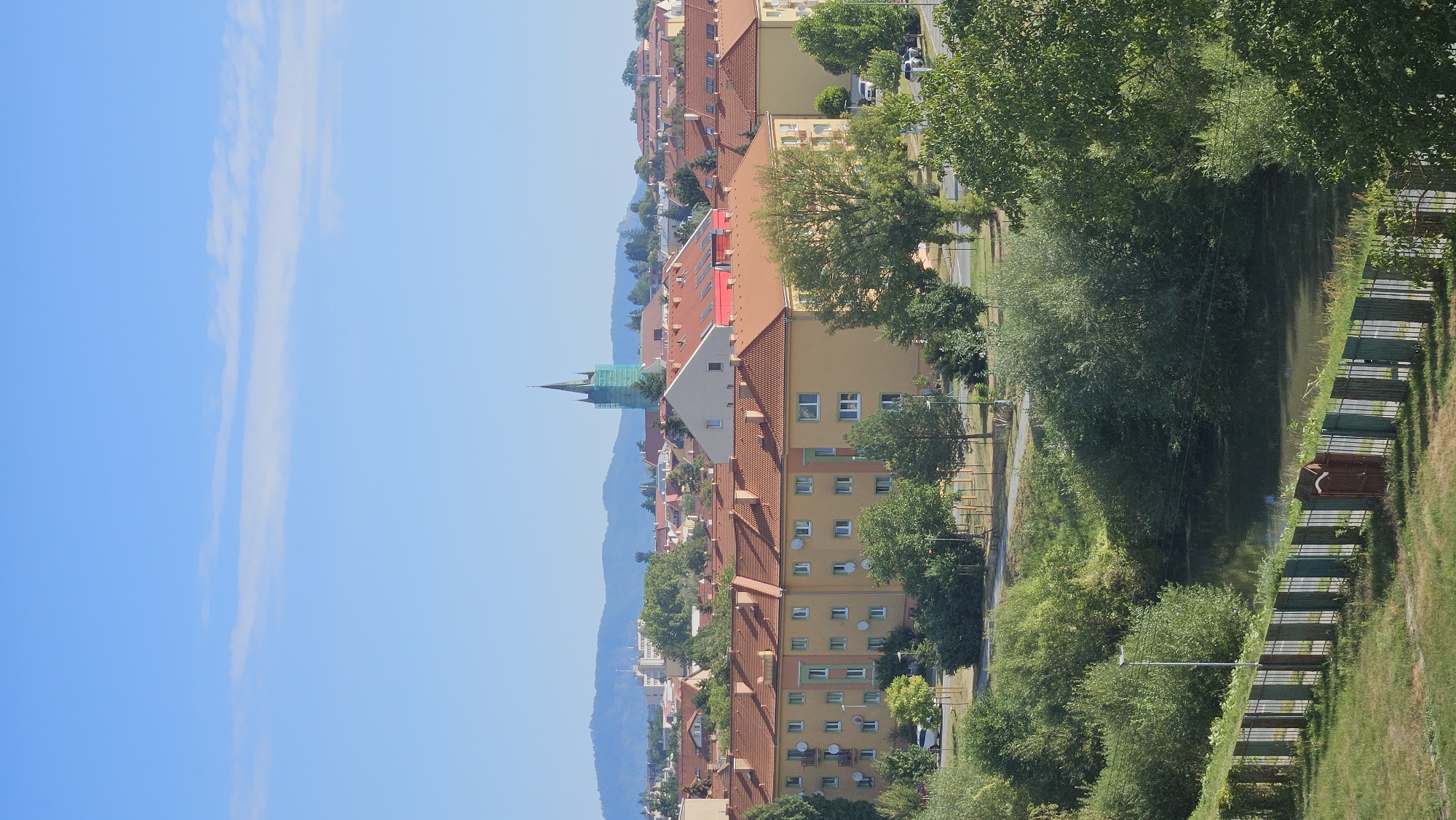 The image depicts a panoramic view of a residential area in Prešov, Slovakia, likely during daytime under a clear sky with some light clouds. In the foreground, a body of water, possibly a river or canal, is visible, bordered by a metal fence. Lush green trees and bushes line the water's edge and extend into the midground, partially obscuring the view of buildings.
The midground is dominated by a row of apartment buildings with orange tiled roofs, typical of Eastern European residential architecture. These buildings are painted in light yellow and beige colors, with visible windows and balconies. A street with parked cars runs in front of these buildings, and more trees are interspersed between them.
In the background, the cityscape of Prešov unfolds, characterized by a collection of buildings with red and orange roofs. A prominent church spire, partially covered by scaffolding or construction netting, stands out against the sky. Beyond the city, rolling hills or mountains are visible in the distance, bathed in sunlight. The overall impression is a peaceful urban landscape on a sunny day.