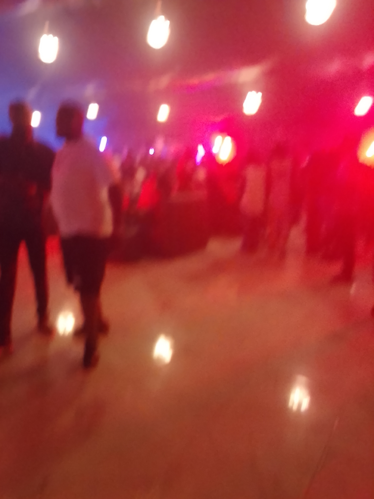 The image captures a blurry, low-light scene of an indoor gathering in Rido, Nigeria. Multiple human figures are visible throughout the frame, largely as indistinct silhouettes or blurred shapes, suggesting movement within a crowd. In the foreground on the left, two figures are somewhat discernible; one on the far left appears to wear dark clothing, while another in the center-left has a light-colored top and darker bottoms.
The setting is characterized by vibrant artificial lighting, with a strong presence of red and blue ambient light. Numerous bright, glowing light fixtures, resembling suspended bulbs or lamps, are visible in the upper part of the frame and their reflections can be seen on the light-colored, possibly reflective floor. These lights appear predominantly white or yellow, contrasting with the dominant red and blue hues of the space.
The overall atmosphere suggests a lively social event, such as a party, club, or performance, taking place at night or in a dimly lit indoor environment. The blurriness indicates motion, implying that people are socializing, dancing, or moving through the venue. No visible text is present in the image.