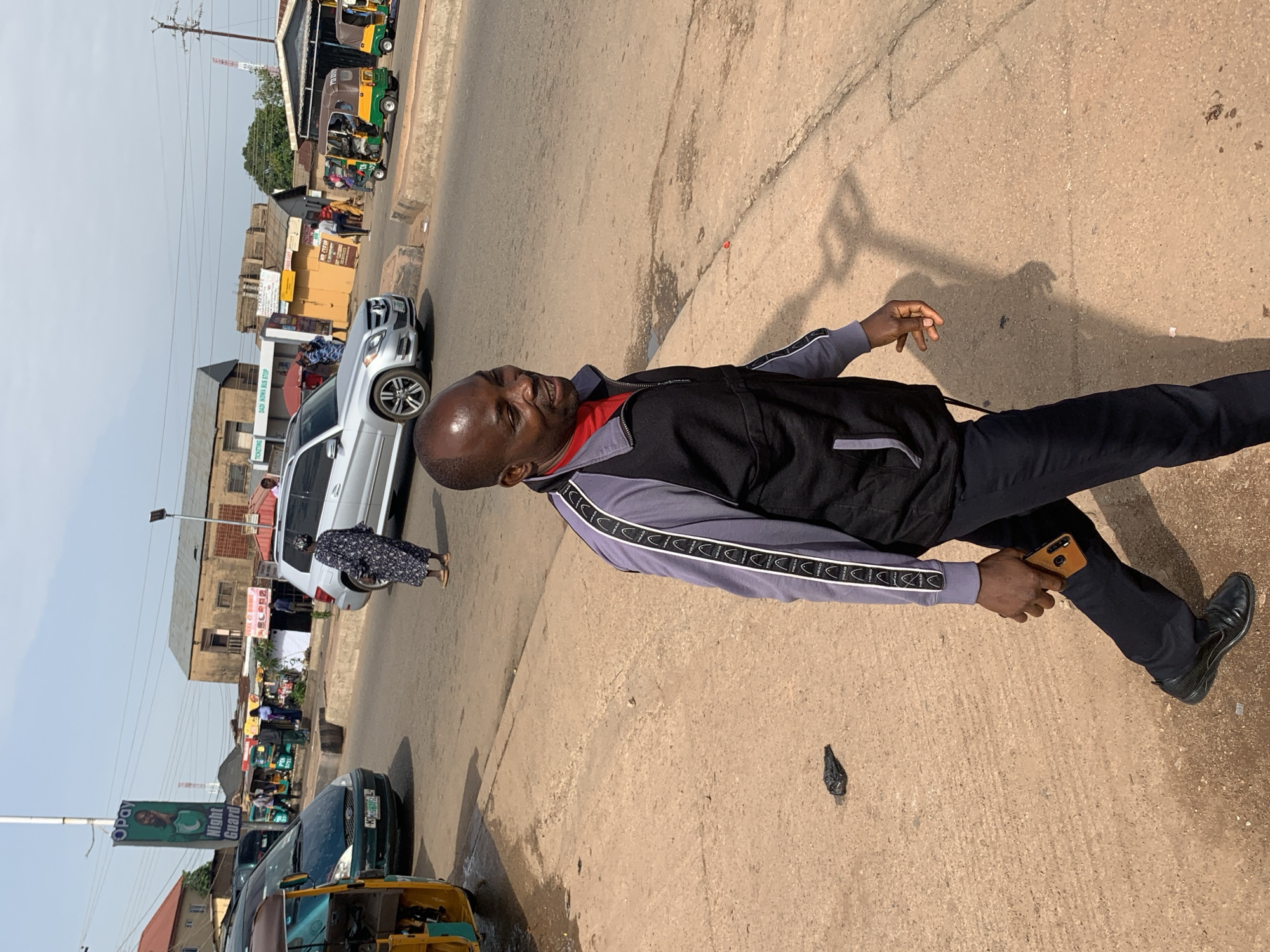 The image captures a street scene in Gura Top, Nigeria, likely during daytime with clear skies. In the foreground, a dark-skinned man is walking towards the right of the frame. He is wearing a black zip-up jacket over a red shirt and dark trousers. He is holding a smartphone in his right hand, which is tucked into his trouser pocket. His left hand is slightly raised, with fingers splayed. His gaze is directed forward and to the right, and he appears to be smiling.
The man is walking on a dirt and asphalt pavement. Behind him, the street is visible with several vehicles, including a silver Mercedes-Benz SUV, a dark green passenger car, and multiple yellow and green three-wheeled auto-rickshaws (known locally as Keke Napep). A woman wearing a patterned blue and white dress is crossing the street in the background, near the SUV.
Further back, buildings and street stalls line the road. A large green billboard for "OPay Night Guard" is prominently displayed on a pole. The environment appears to be a busy urban area with signs of commercial activity and public transportation. The ground is dusty and shows some signs of wear and tear.