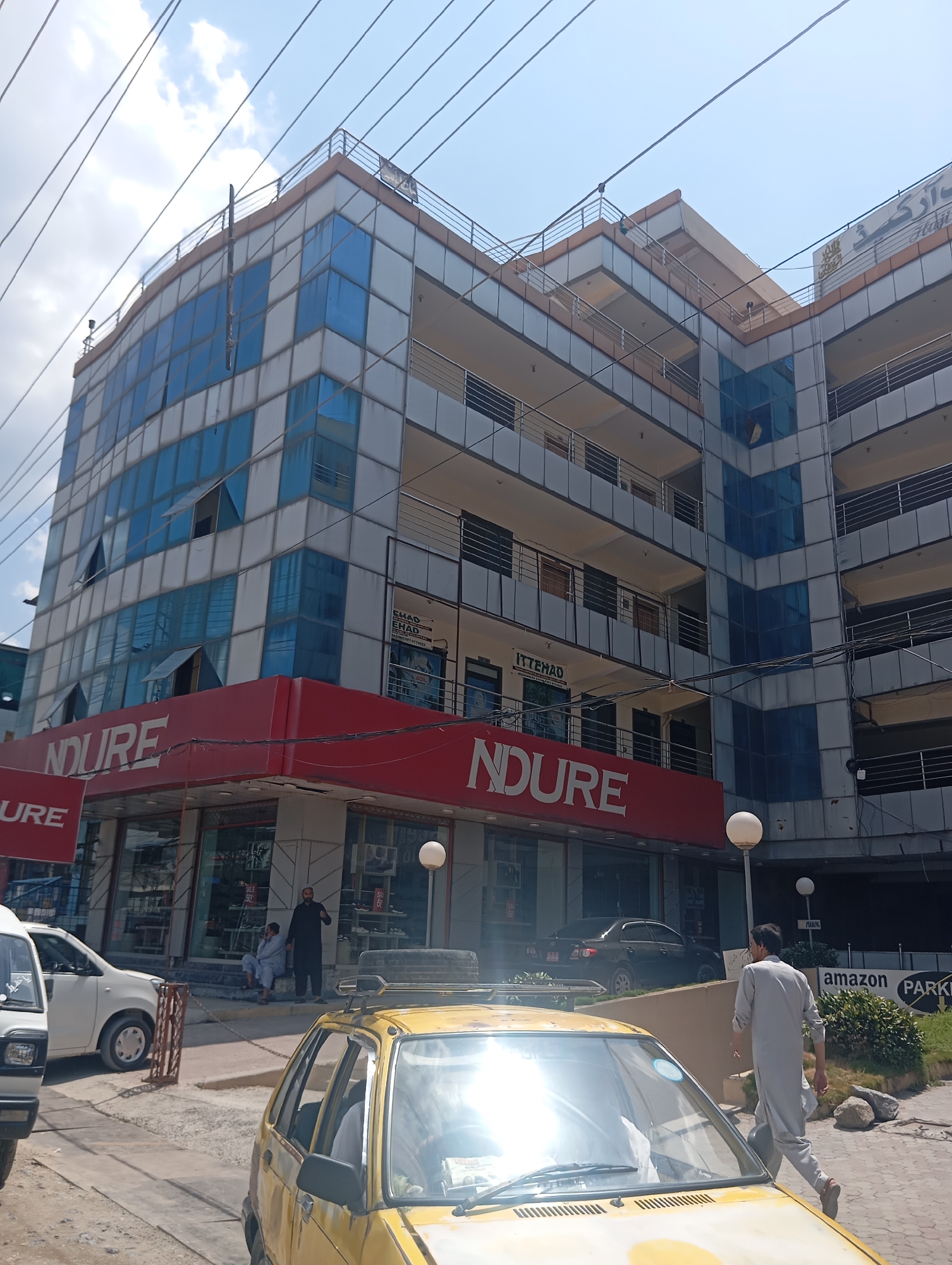 A multi-story building with a modern facade featuring blue and white glass panels dominates the scene, with balconies on several levels. A red awning stretches across the ground floor, displaying the brand "NDURE" in large white letters. Below the awning, storefronts are visible with displays of merchandise and signage, including "SALE 50%". A white car is parked to the left of the building, and a yellow car is in the foreground on the right. Two men are standing near the entrance of the building, one in a black shalwar kameez and the other in a light blue shalwar kameez, appearing to be in conversation. Another man in a light grey shalwar kameez walks towards the right side of the image, carrying a bag. A sign for "amazon PARKING" is visible in the background. Power lines are strung across the sky, which is partly cloudy. The overall impression is a commercial area on a bright day.
