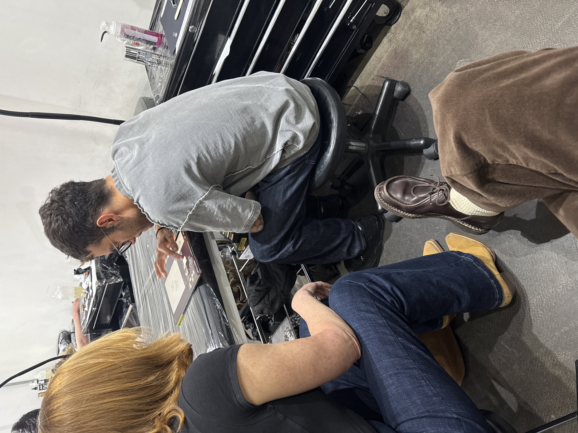 The image shows three people gathered around a table. A man wearing glasses and a gray shirt sits on a rolling chair, looking at a tablet. A person with blonde hair is wearing a black shirt and seated in front of the man with glasses. Another person is seated to the side, wearing brown pants and brown shoes with white socks. The scene is likely indoors, possibly in a shop, with a black tool cabinet visible in the background. The location in London, United Kingdom, cannot be determined from the provided image.
