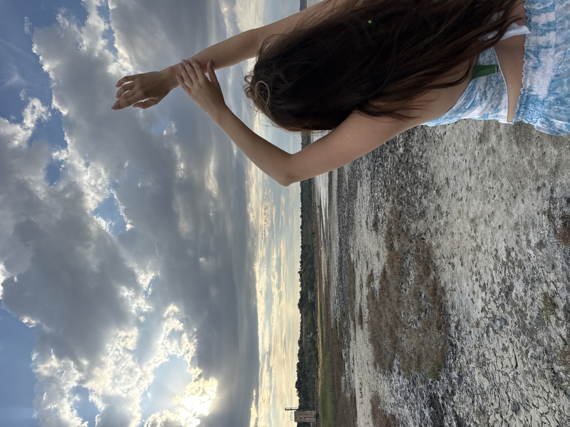 The media file depicts a young woman with long, dark hair in a blue and white dress, captured from behind as she looks up at a dramatic sky. Her arms are raised, with one hand resting on her wrist, suggesting a pose of contemplation or stretching. The scene appears to be outdoors, with a cracked, dry ground underfoot and a distant, hazy landscape. The sky is filled with a mix of dark, heavy clouds and brighter patches where sunlight breaks through, indicating a dynamic weather condition, possibly a transition from rain or an approaching storm. The time of day seems to be late afternoon or early evening, given the quality of the light. A small, rustic-looking structure is visible in the distance on the horizon. The overall mood conveyed is one of solitude and awe, with the vastness of the sky juxtaposed against the stillness of the woman and the arid environment. The location is Odesa, Ukraine. There is no visible text in the media file.