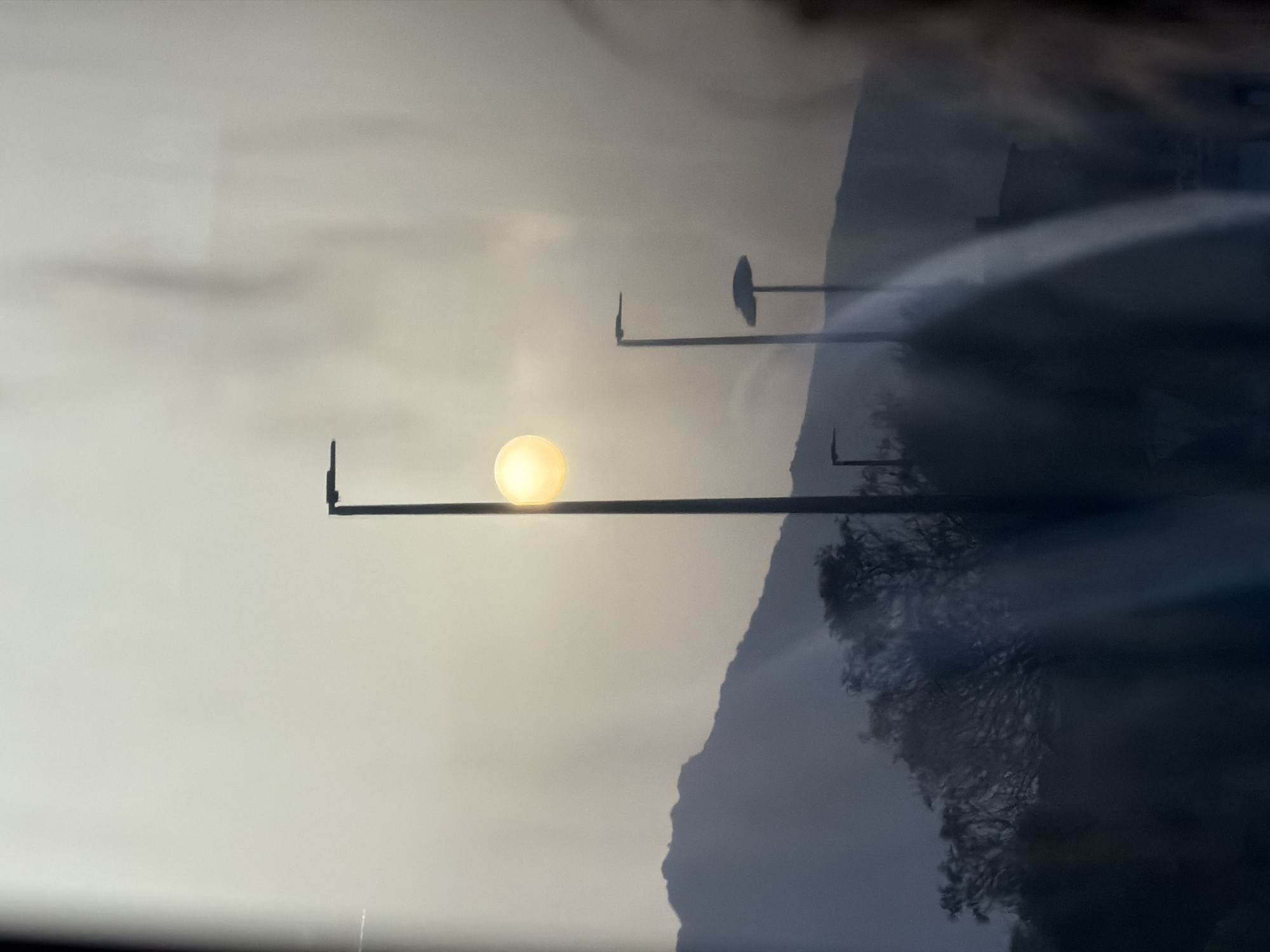 This image depicts a hazy daytime scene with the sun prominently visible through atmospheric conditions. The sun, a bright yellow orb, is partially obscured by a light mist or haze, creating a soft glow. It is positioned behind and slightly to the right of a tall, dark lamppost.

In the background, a silhouetted mountain range stretches across the horizon. The mountains are dark and appear to have a jagged, uneven skyline. In the foreground, several other lampposts are visible. One is located to the right of the central lamppost and appears shorter. Further to the left, a third lamppost is barely visible against the hazy sky.

The foreground also contains dark, out-of-focus shapes that suggest trees and possibly buildings. The overall impression is one of a landscape seen through a window or from a distance, with a slight reflection or distortion on the surface. There are no people clearly visible in the image. The scene conveys a sense of quietness and diffused light.