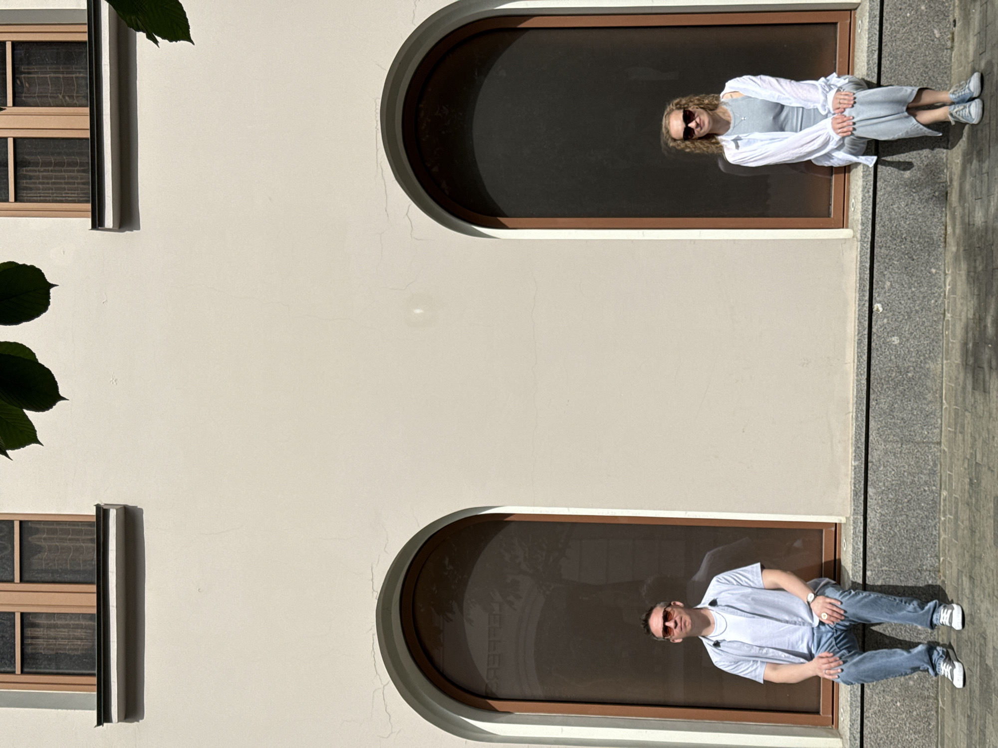 A rotated, high-angle shot depicts a light-beige building facade featuring two arched window recesses. Each recess contains a person sitting. The upper window features a light-haired woman wearing sunglasses, a grey dress, and a white shirt. The lower window features a fair-skinned man wearing sunglasses, a white t-shirt, and blue jeans. Both appear to be on a sidewalk directly in front of the building. Leaves are visible on the upper left side of the frame. Two rectangular windows are on the left side of the frame. The city is Moscow, Russia.