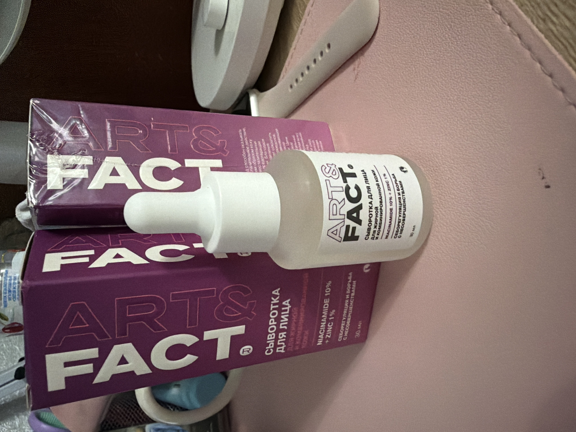 The image displays two boxes of "ART&FACT" serum for the face, alongside a bottle of the same product. One box is partially visible on the left, and another is to its right. The bottle, featuring a white dropper top, is positioned in front of the boxes. Text on the packaging, visible in both Russian and English, indicates the product is a serum for oily and combination skin, containing Niacinamide 10% and Zinc 1%. The scene is set indoors on a pink textured surface, possibly a desk mat, with a white watch band visible to the right. The background includes a white charging device and other indistinct objects. There is no indication of the city of Chaikovsky, Russia, or any geographical markers.