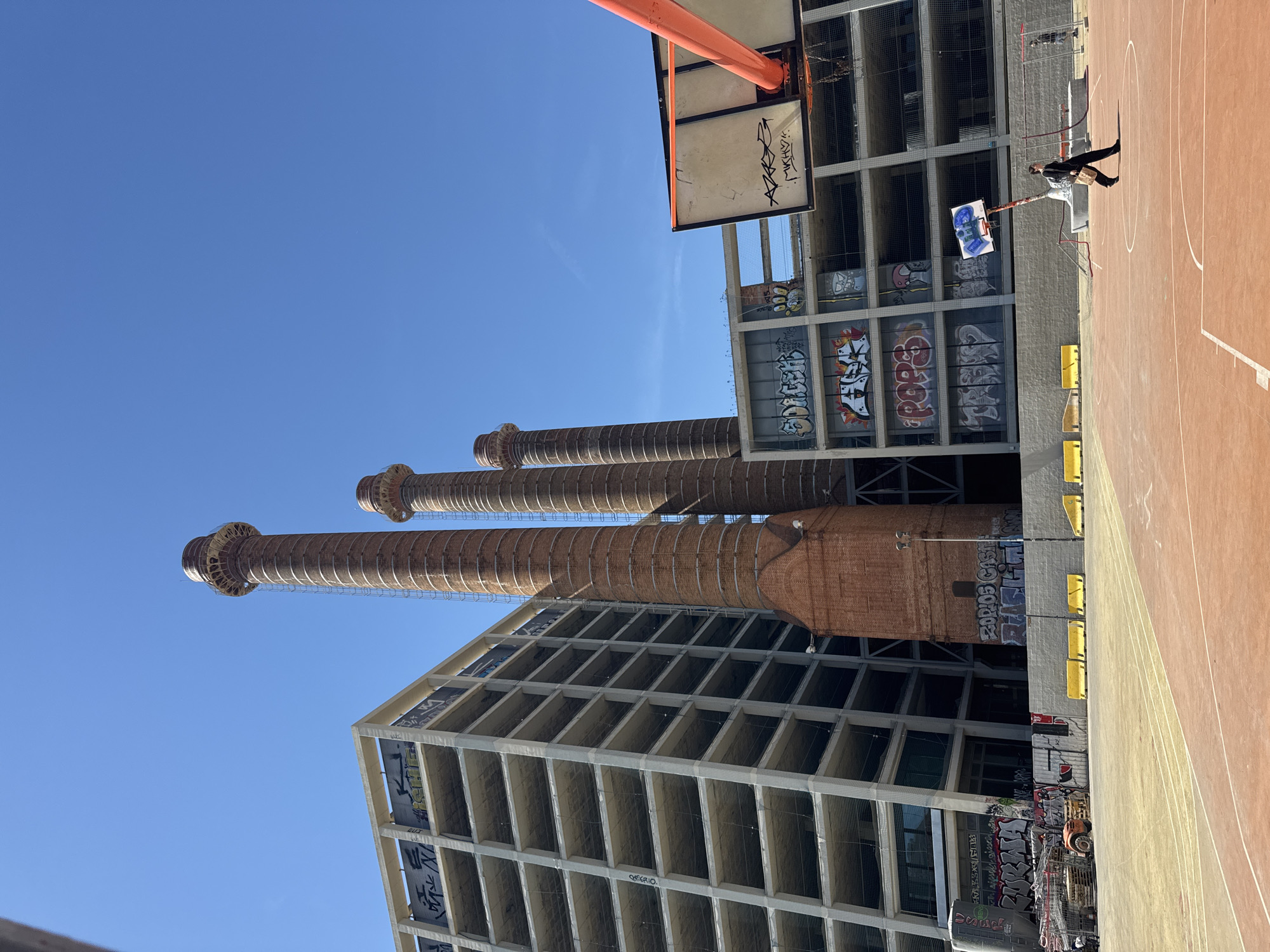 The image captures an urban scene featuring prominent industrial architecture juxtaposed with a recreational space under a clear blue sky.
In the background, three towering brick industrial chimneys stand tall against the bright sky, displaying aged texture and intricate tops. Adjacent to them, and extending into the midground, is a multi-story, unfinished or derelict concrete building characterized by numerous open, grid-like cells. The walls of this building are extensively covered in various graffiti tags and murals.
In the foreground and midground, a reddish-orange basketball court with white markings is visible. A single basketball hoop with an orange rim and a white backboard is present, the latter also bearing black graffiti. One person is standing on the basketball court, positioned near the hoop. They appear to be wearing dark clothing, possibly a jacket and pants, and are carrying a bag over their shoulder. Along the edge of the court, several yellow barriers or blocks are visible, and further back, some utility or construction equipment can be seen near the building.
The scene takes place on a clear, sunny day, indicated by the vibrant blue sky and distinct shadows cast by the structures and the person. The overall setting is an urban environment, likely a revitalized or repurposed industrial area.
Visible text includes numerous graffiti tags on the building walls and the basketball backboard. Examples on the building include "2DUDOS," "QHEORIO," "POPOS," "CASA," and "SEK," among many others that are highly stylized or partially obscured. The backboard graffiti is illegible but appears to be a stylized word.