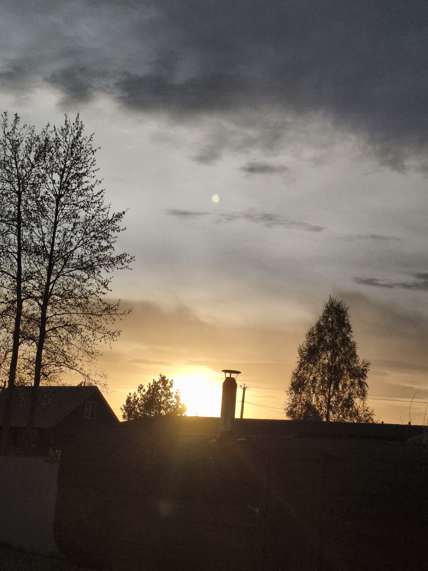 The image shows a sunset in Русский Вожой, Russia. The sky is filled with dark gray clouds above a layer of orange and yellow light from the setting sun. Silhouetted in the foreground are trees, the roof of a building, and a chimney. An overhead power line is visible between the chimney and one of the trees. The lower part of the image is dark, obscuring details of the ground.