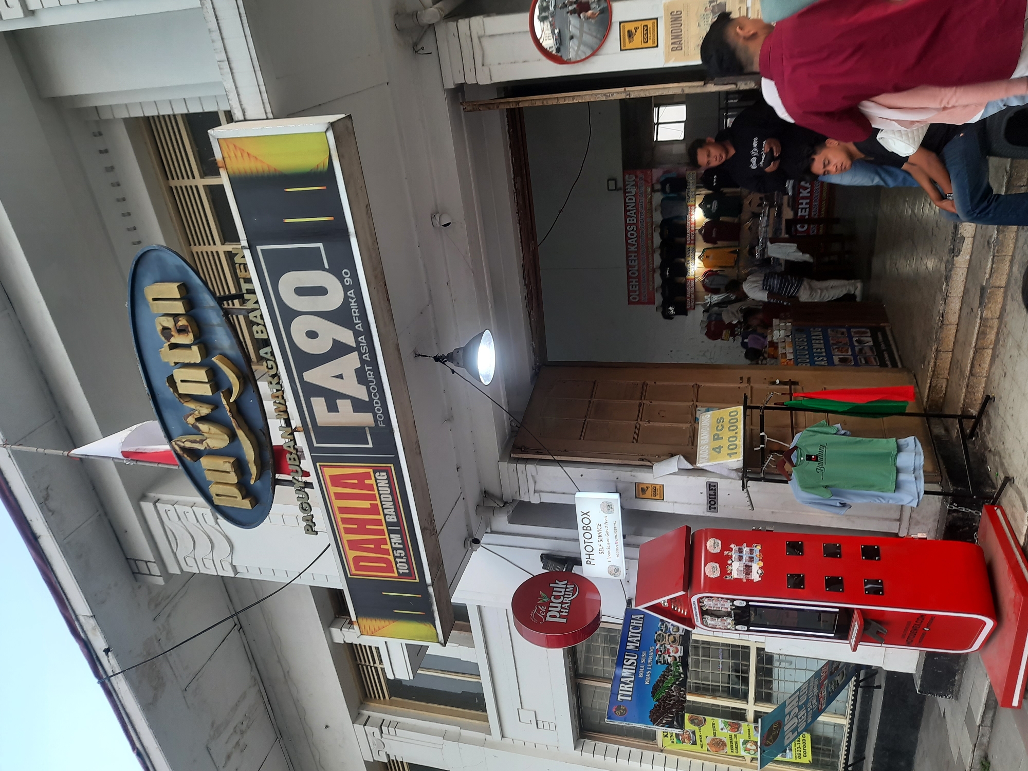 The image shows a street view in Bandung City, Indonesia, featuring a store with various signs and products. Prominently displayed is a large blue and gold oval sign that reads "Puwn ten" with "PAGUYUBAN WARGA BANTEN" underneath. Below this, a rectangular sign features "DAHLIA 101.5 FM BANDUNG" in red and yellow, next to another sign with "FA90 FOODCOURT ASIA AFRIKA 90" in large white letters on a black background.
To the left, a red, house-shaped machine is positioned on the sidewalk, likely a photo booth or vending machine, with the brand "MOTODEWA.COM" visible at its base. Nearby, a circular red sign advertises "Teh Pucuk Harum". A sign for "TIRAMISU MATCHA" with images of food is also visible.
Inside the open storefront, shelves are stocked with clothing, including t-shirts displayed on racks. A sign above reads "OLEH OLEH KAOS BANDUNG" (Souvenirs Bandung T-shirts), and further down, a yellow sign with red text states "BANDUNG 4 PCS 100.000".
Several people are present in the scene. Two individuals are seated in front of the store, one in blue jeans and a black shirt, the other wearing a maroon shirt. Another person is standing behind them. A person wearing a striped shirt and white pants is seen walking into the store. Other individuals are partially visible within the store's interior.
The environment appears to be an urban street with a sidewalk and the facade of a building, suggesting a commercial area. The lighting indicates it is either early morning or late afternoon.