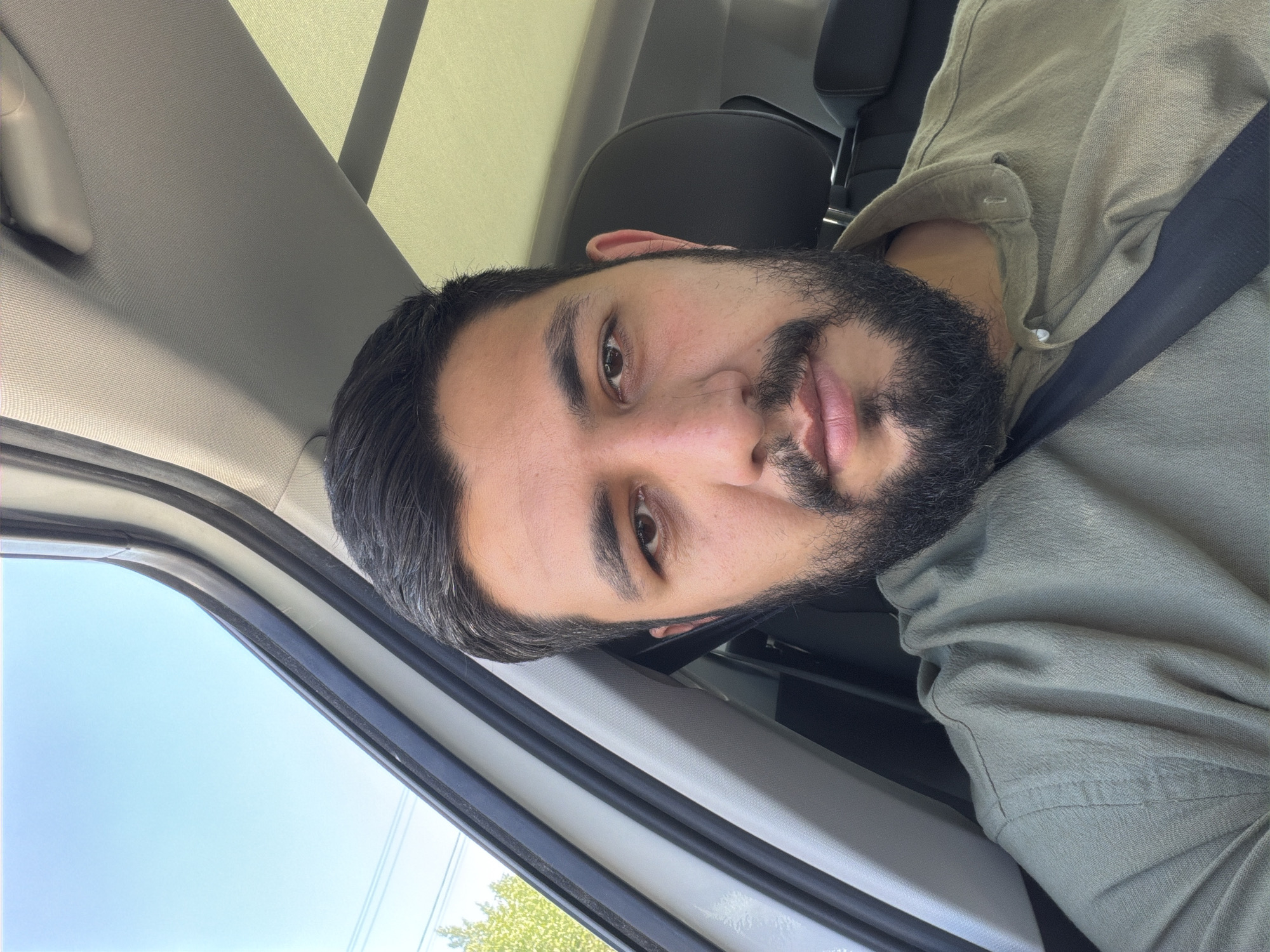 The image captures a man in the interior of a car, looking directly at the camera. He has dark hair, a full beard, and a mustache, with dark eyes. He is wearing a light olive-green or khaki-colored collared shirt, and a dark seatbelt is visible diagonally across his chest.
The setting is clearly inside a vehicle. Visible car components include a light grey headliner, a sun visor, and a dark car seat behind the man. Through the car window on the left side of the frame, a clear, bright blue sky is visible, indicating daytime. Below the sky, some green foliage from a tree is partially seen.
The man maintains a neutral expression. The overall lighting is bright and natural, consistent with sunny weather. The activity appears to be a casual portrait or selfie taken while inside the car. There is no visible text in the image.
