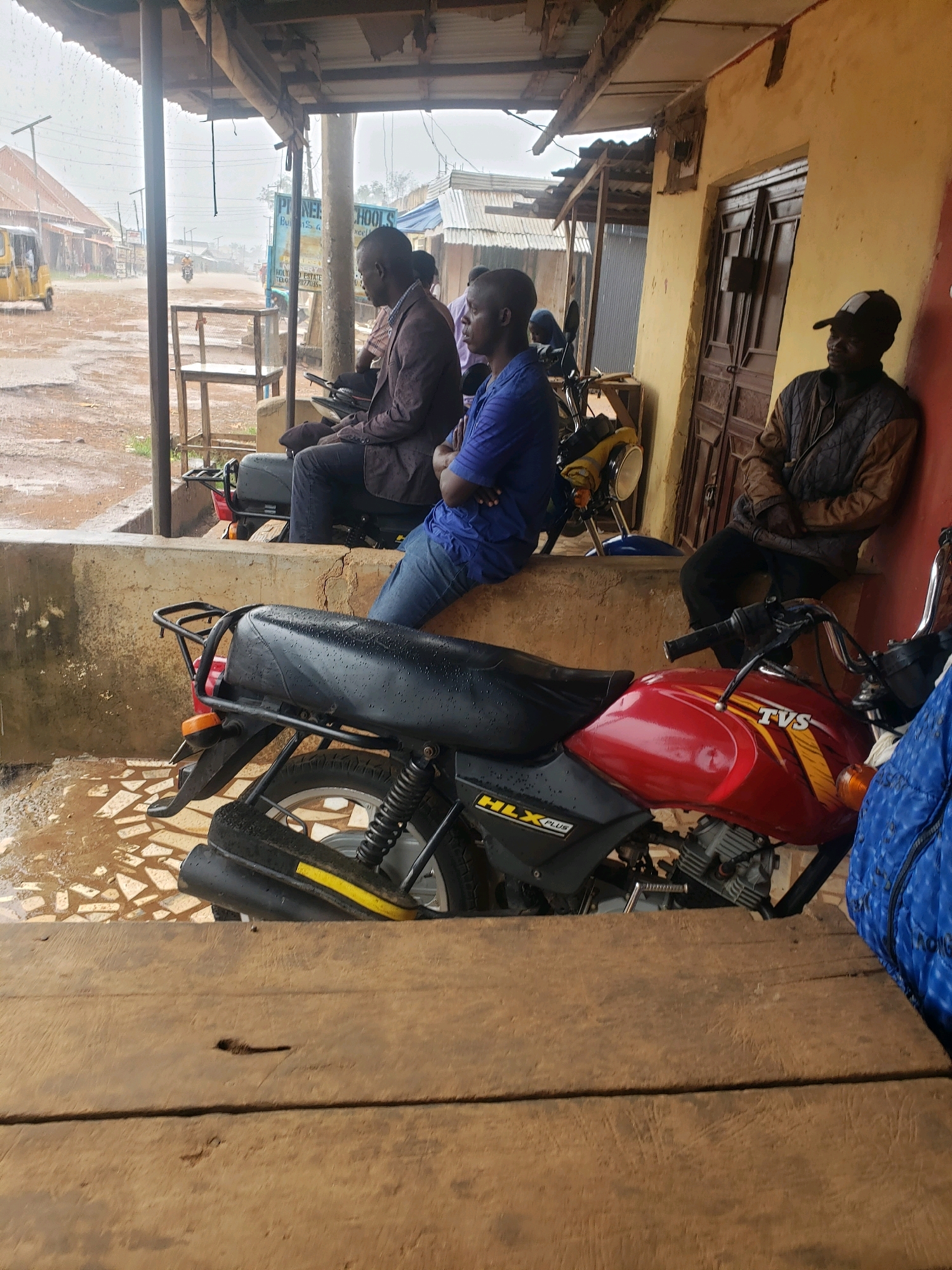 A group of men are gathered under a covered outdoor area in Sabon Tasha, Nigeria, on a rainy day. The ground is wet and reflects the overcast sky and surrounding structures. In the foreground, a red and black motorcycle is parked. Three men are visible under the shelter. One man in a dark suit and jeans sits on another motorcycle, looking to his left. Another man in a blue shirt and jeans is seated, facing away from the camera. A third man in a brown jacket and a baseball cap leans against a yellow wall, looking to his right. The background shows a street with buildings, some with corrugated metal roofs, and a yellow auto-rickshaw driving away. The overall scene suggests a brief respite from the rain.
