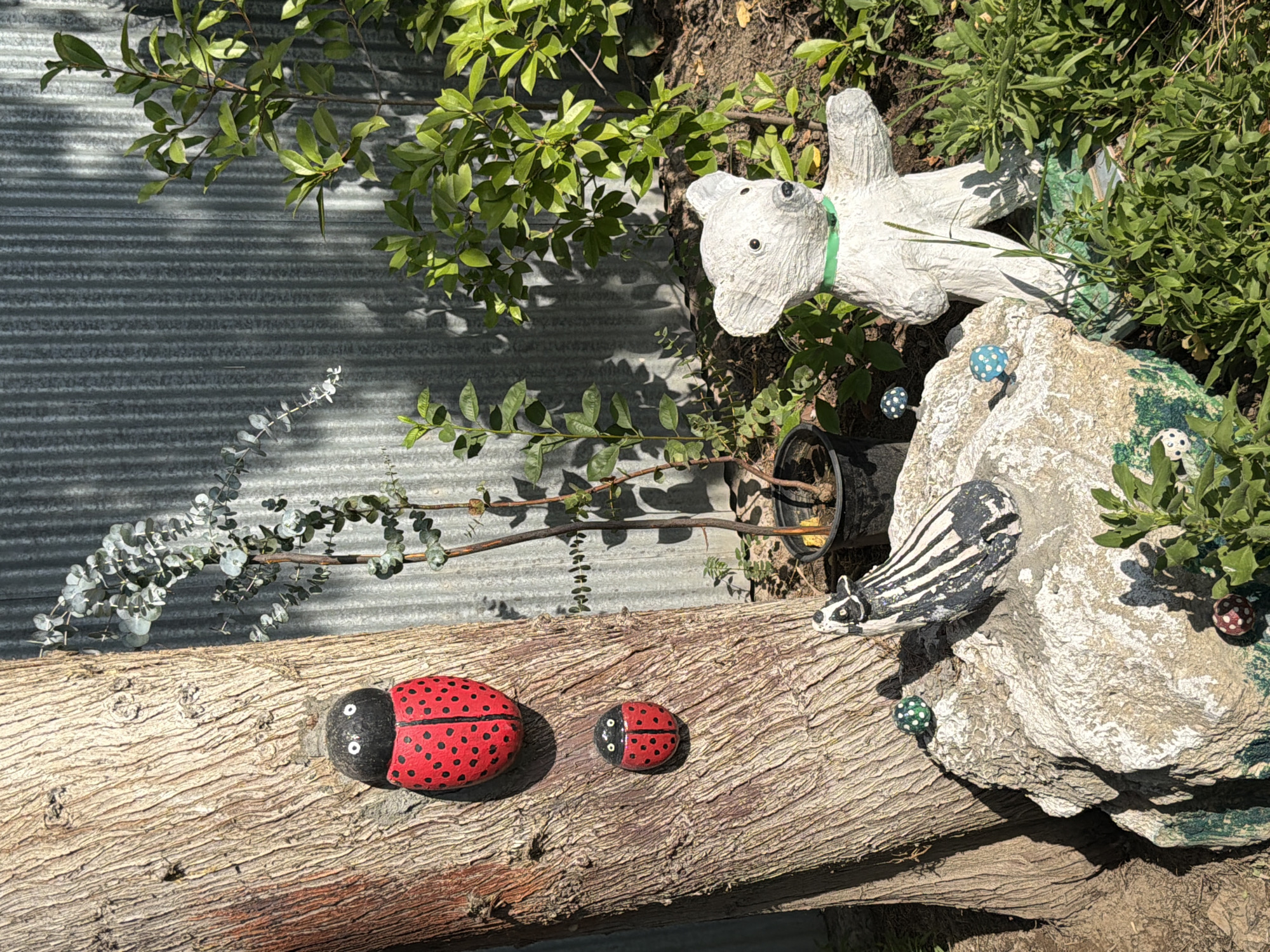This is a close-up, eye-level outdoor shot of a garden scene. In the foreground, a large, textured tree trunk dominates the left side of the frame. Two ladybug decorations, painted red with black spots and black heads with white eyes, are attached to the trunk. The upper ladybug is larger than the lower one. To the right of the tree, a large grey corrugated metal fence forms the background. In front of the fence, a potted eucalyptus plant with sparse, grey-green leaves is visible. To the right of the eucalyptus, a white bear statue with a green collar stands among lush green foliage. In the mid-ground, a rough, light-colored rock is situated in front of the tree trunk. On the rock, a black and white striped animal figurine, possibly a skunk or badger, is placed. Several small, decorative mushrooms with white caps and blue or red spots are scattered around the rock. The overall lighting suggests a sunny day, casting shadows from the plants onto the fence. The location is identified as Balakənd, Azerbaijan.