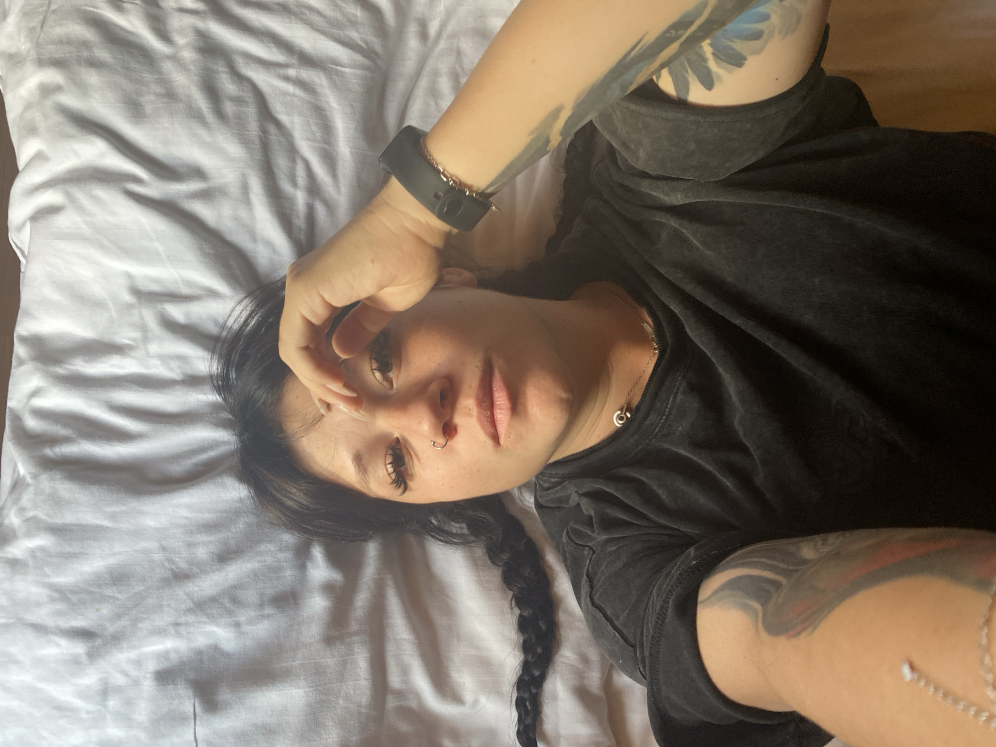 The image shows a woman lying on a white, wrinkled sheet, likely a bed. She has dark hair styled in two braids, one of which extends to her left. Her right hand is resting on her forehead, and she is wearing a dark t-shirt. She has a nose ring and what appears to be extensive tattoos on her arms. A dark smartwatch is visible on her left wrist. The lighting suggests an indoor setting. The image does not contain any elements that indicate a specific location within Krasnoflotskoe, Russia.