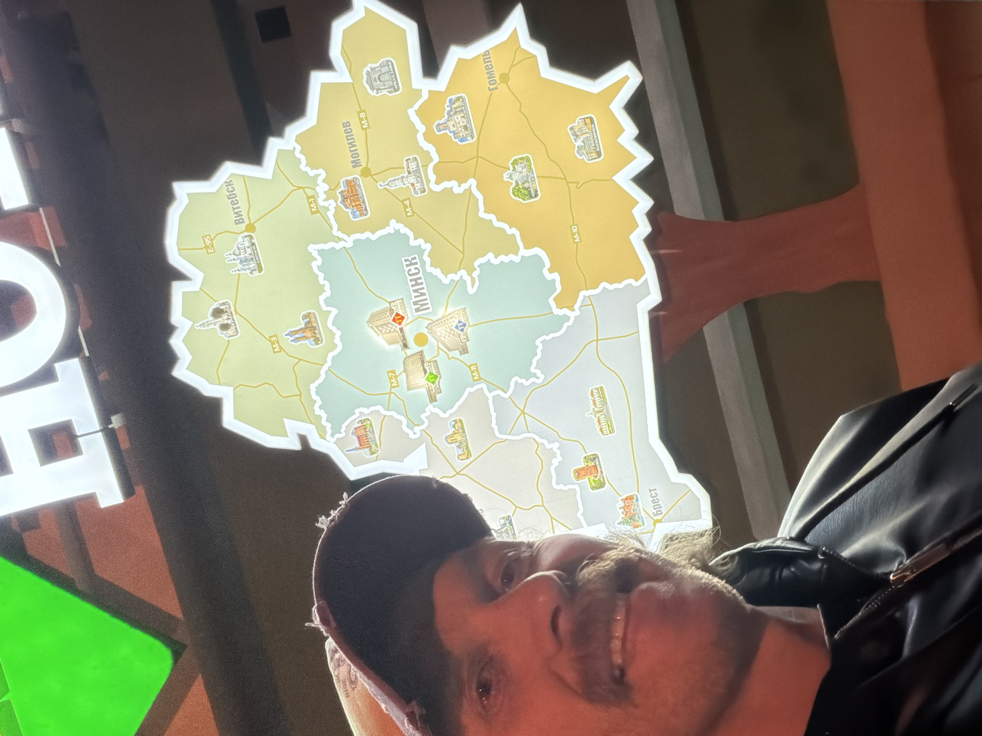 The image shows a smiling man in a dark cap and black leather jacket, appearing to take a selfie or be photographed in front of a brightly lit, stylized map of Belarus. The map is divided into its six administrative regions (oblasts), each depicted with a distinct pastel color and featuring illustrations of local landmarks or prominent buildings.
The central region is labeled "МИНСК" (Minsk), the capital. Other visible city labels include "Витебск" (Vitebsk) in the north, "Могилев" (Mogilev) in the east, "Гомель" (Gomel) in the southeast, and "Брест" (Brest) in the southwest. Major roads and highways are marked with their designations, such as M-1, M-3, M-4, M-7, M-8, M-10, M-11, and E-95. Near Mogilev, the text "АРКА СЛАВЫ" (Arch of Glory) is visible.
The man has a mustache and beard, and his bright smile suggests a positive mood. Partially visible to the left are large, illuminated letters, reading "HO" and "TEL", potentially part of a "HOTEL" sign. The word "FOTEL" is also seen in several places on the map itself. The background is dark, indicating an indoor setting, possibly a travel-themed display, a hotel lobby, or an information center within Minsk, Belarus. The image is captured from a slightly upward angle, with the man's head and shoulders framing the lower part of the map.