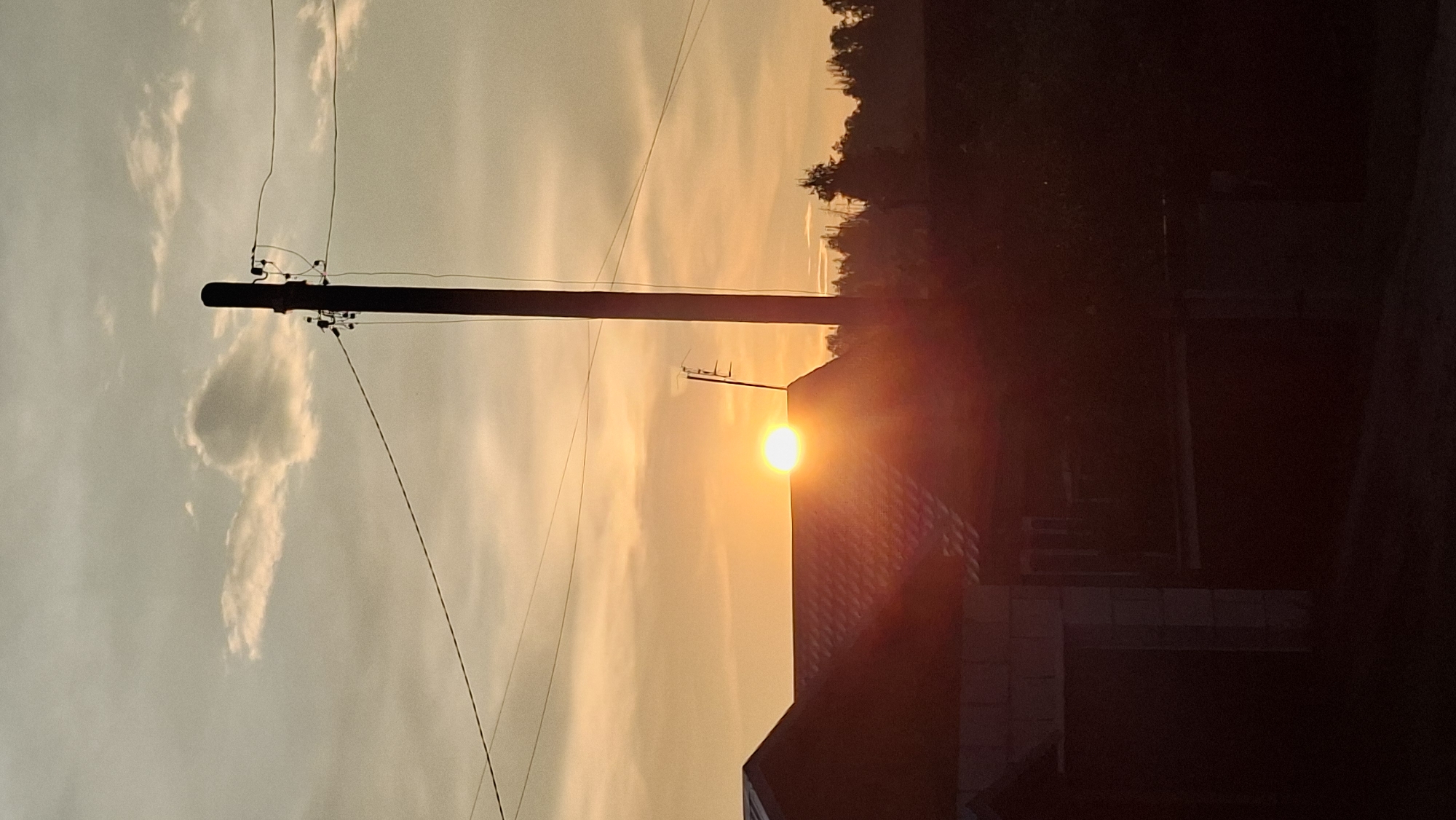 The image captures a sunset over what appears to be a residential area in Novoilinsky, Russia. A prominent utility pole with insulators and wires stands in the center-right of the frame, silhouetted against the bright orange sky. The sun, a glowing orb, is setting behind the rooftop of a dark, silhouetted house in the foreground, casting a strong lens flare. The roof of the house is tiled and appears to have a slight sheen from the setting sun. Beyond the house, a line of trees forms a dark, silhouetted treeline against the horizon. The sky is filled with soft, diffused clouds, tinged with the warm hues of sunset. The overall impression is one of a rural or semi-rural landscape bathed in the warm light of dusk.