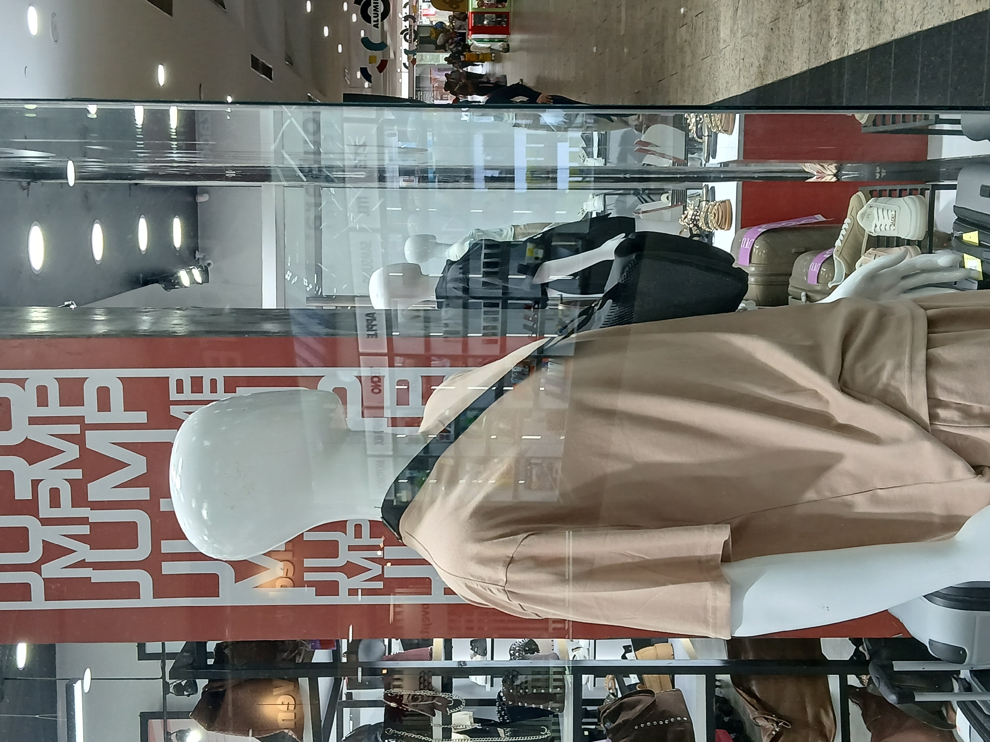 The image shows a display case in a store. The display features a mannequin wearing a tan-colored outfit. Behind the mannequin, there's a red panel with white lettering that appears to say "JUMP". The reflection in the display case shows the interior of a shopping mall with people walking and various stores visible in the background. There are lights on the ceiling of the mall. The lower shelves of the display case contain handbags and other accessories. This is likely a store located in a shopping mall in Ciudad Guayana, Venezuela.