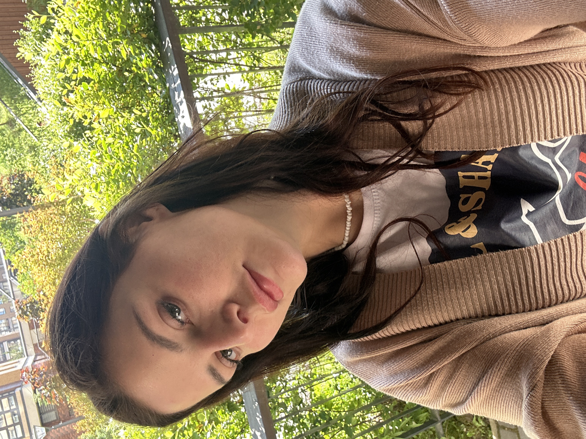 A woman is photographed from the chest up, looking at the camera with a slight smile. She has long, dark brown hair that falls over her shoulders. She is wearing a beige cardigan over a white t-shirt with a graphic print. A pearl necklace is visible around her neck. The background is out of focus, showing lush green foliage and hints of buildings. The lighting suggests it is daytime, possibly outdoors on a balcony or patio.