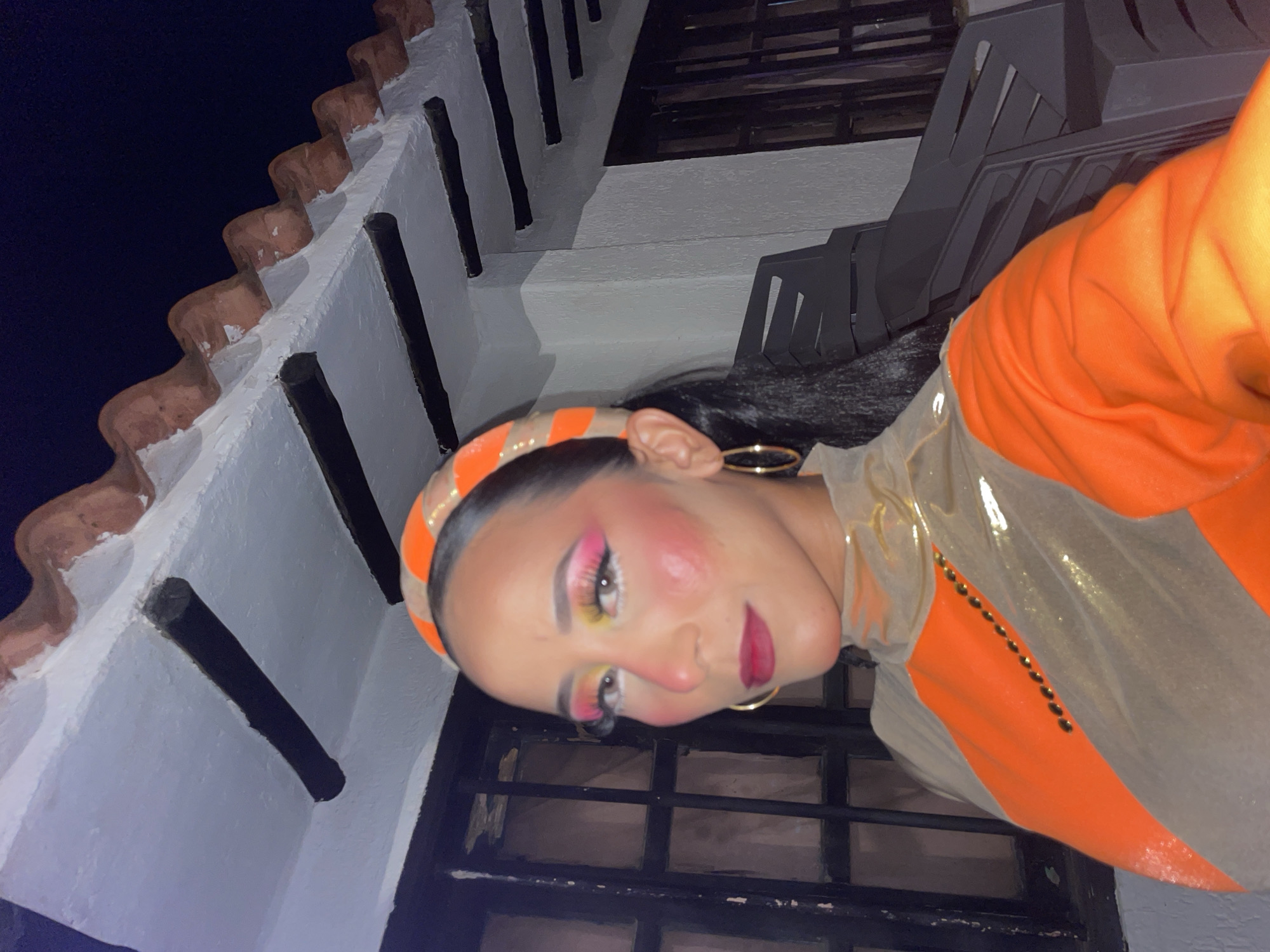 A medium shot captures a woman with dramatic makeup and an orange and beige outfit. She wears bright pink and yellow eyeshadow, thick eyeliner, long false eyelashes, and bold pink lipstick. Her cheeks are heavily rouged. A striped orange and beige headband adorns her dark hair, which is pulled back into a ponytail. She has large gold hoop earrings. The outfit features a high neckline with sheer, shimmery beige fabric draped over an orange garment. A line of small black beads is visible on the chest area. She is positioned in front of a building with a white facade and dark window frames, possibly a home or establishment. Above, a rustic roof with terracotta tiles is visible against a dark night sky. The overall scene appears to be outdoors, likely in a warm climate, given the architecture. The lighting suggests it is evening or night. This setting is consistent with the architectural style found in Porlamar, Venezuela.