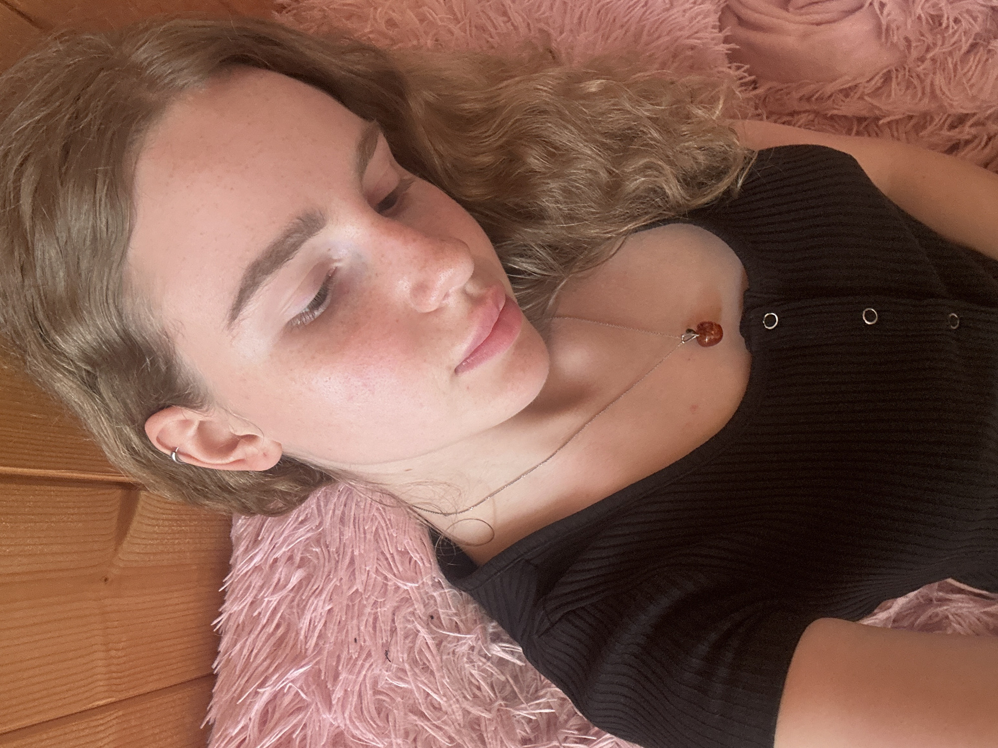 A young woman is captured in a close-up, high-angle shot, seemingly resting or sleeping. Her eyes are closed, and her lips are slightly parted, conveying a peaceful and relaxed expression. She has light brown, wavy hair that flows around her head and shoulders. Her fair skin shows a scattering of faint freckles across her nose and cheeks.
She is wearing a fitted black top with a ribbed texture, featuring three small, silver-ringed buttons or snaps on the upper chest area. A delicate silver chain necklace with a small, reddish-brown, translucent pendant rests on her skin. A simple silver hoop earring is visible in the helix of her left ear.
The woman is lying on a plush, fluffy pink blanket or throw, which occupies much of the background. To the left of the frame, warm-toned wooden paneling is visible, suggesting an indoor setting with wooden walls. The lighting is soft and natural, contributing to the tranquil mood. There is no visible text in the image. The scene suggests a moment of quiet repose in Vasilyevo, Russia.