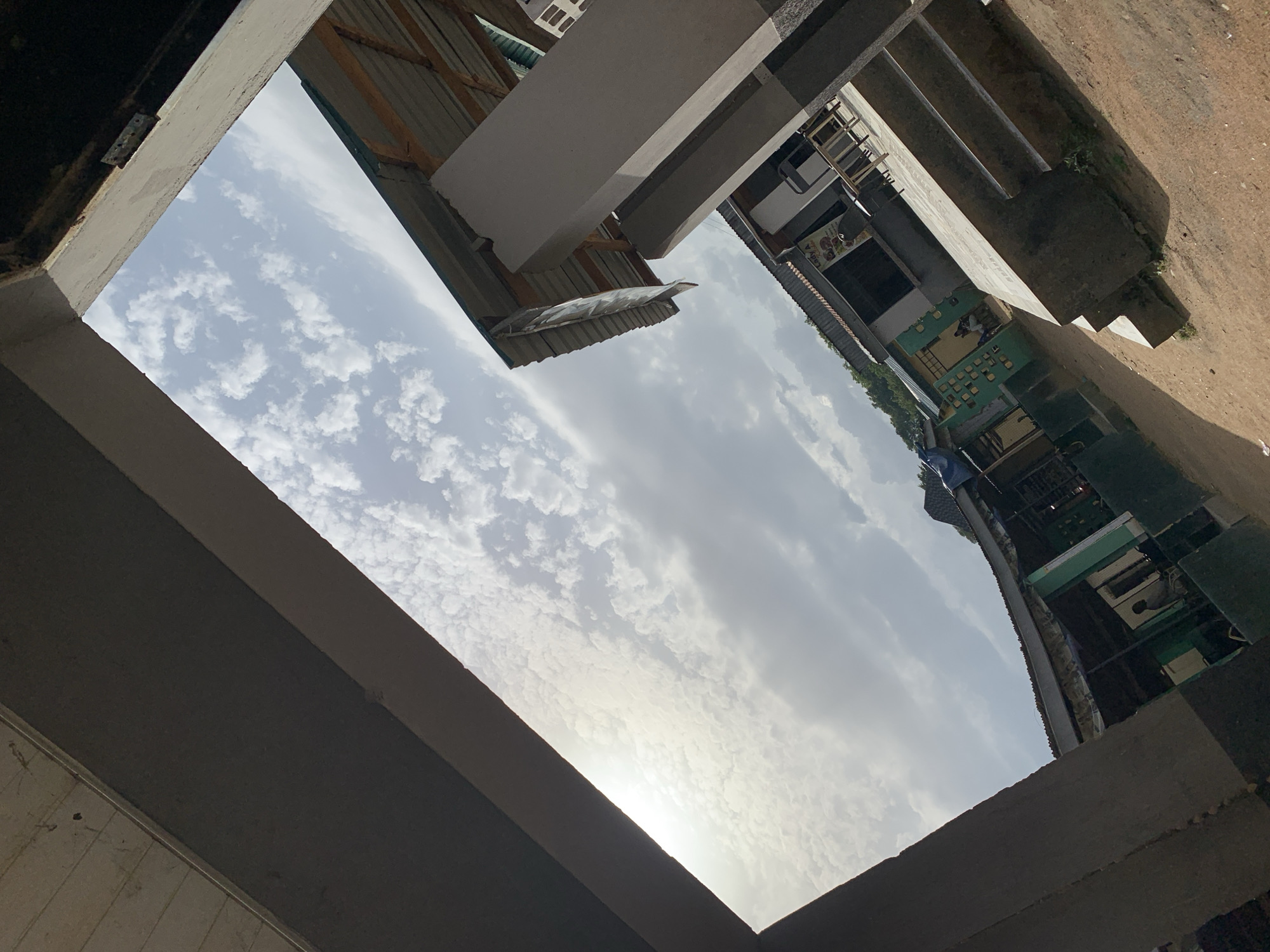 The image depicts an outdoor scene, likely in Jos, Nigeria, viewed from a low angle looking upwards through an opening formed by concrete structures and corrugated metal roofing. The sky is filled with scattered white clouds against a pale blue backdrop, suggesting it is daytime, possibly late morning or early afternoon. Sunlight is visible as a bright, diffused glow towards the left side of the frame, peeking through the clouds.
The foreground and immediate surroundings consist of unfinished or weathered concrete structures, including a large, dark concrete beam dominating the bottom left and a lighter concrete element arching across the top. These structures frame an opening that looks out onto a more distant scene.
In the background, various buildings and structures are visible, characterized by corrugated metal roofs and light-colored walls, some with visible wooden supports. One building appears to have multiple levels and windows. A green-painted section of a building is prominent, with several smaller windows or compartments. Some signage is visible on the buildings, but the text is too small and indistinct to be read. There are also hints of vegetation and what appears to be a dirt or gravel path in the lower right portion of the image.
No specific activity or event is clearly taking place. The scene suggests an urban or semi-urban environment, possibly an area undergoing construction or with informal housing. The overall impression is one of a modest, functional setting bathed in natural light.